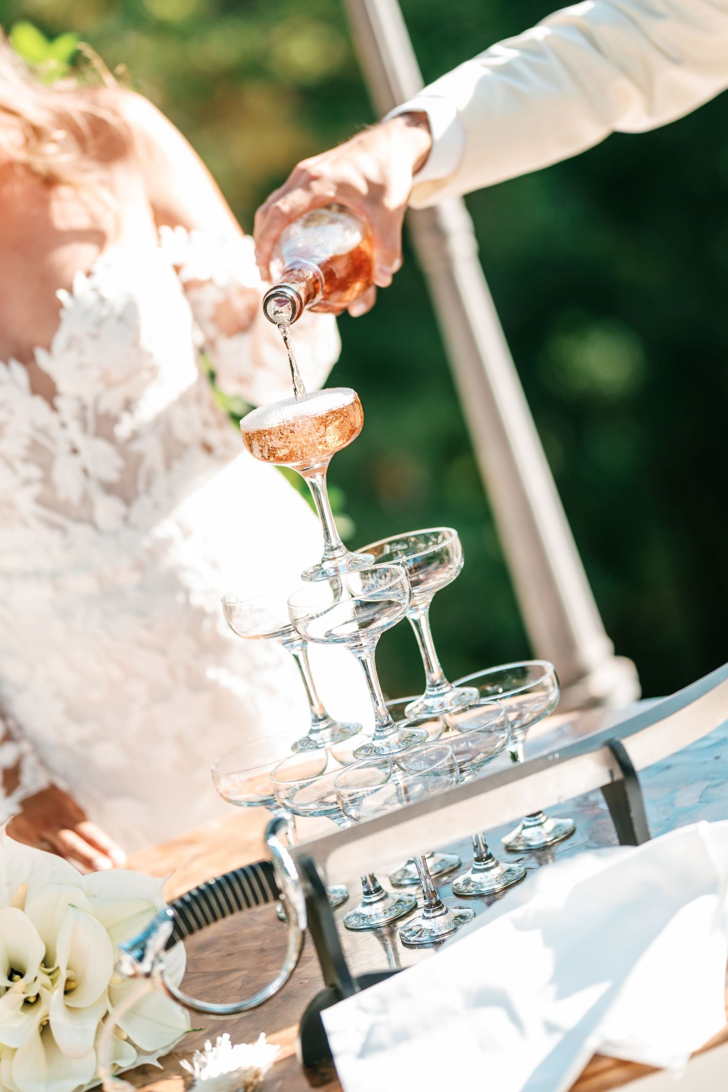 Bride in a lace gown watches as rosé champagne is poured over a tiered champagne tower at an outdoor wedding celebration.