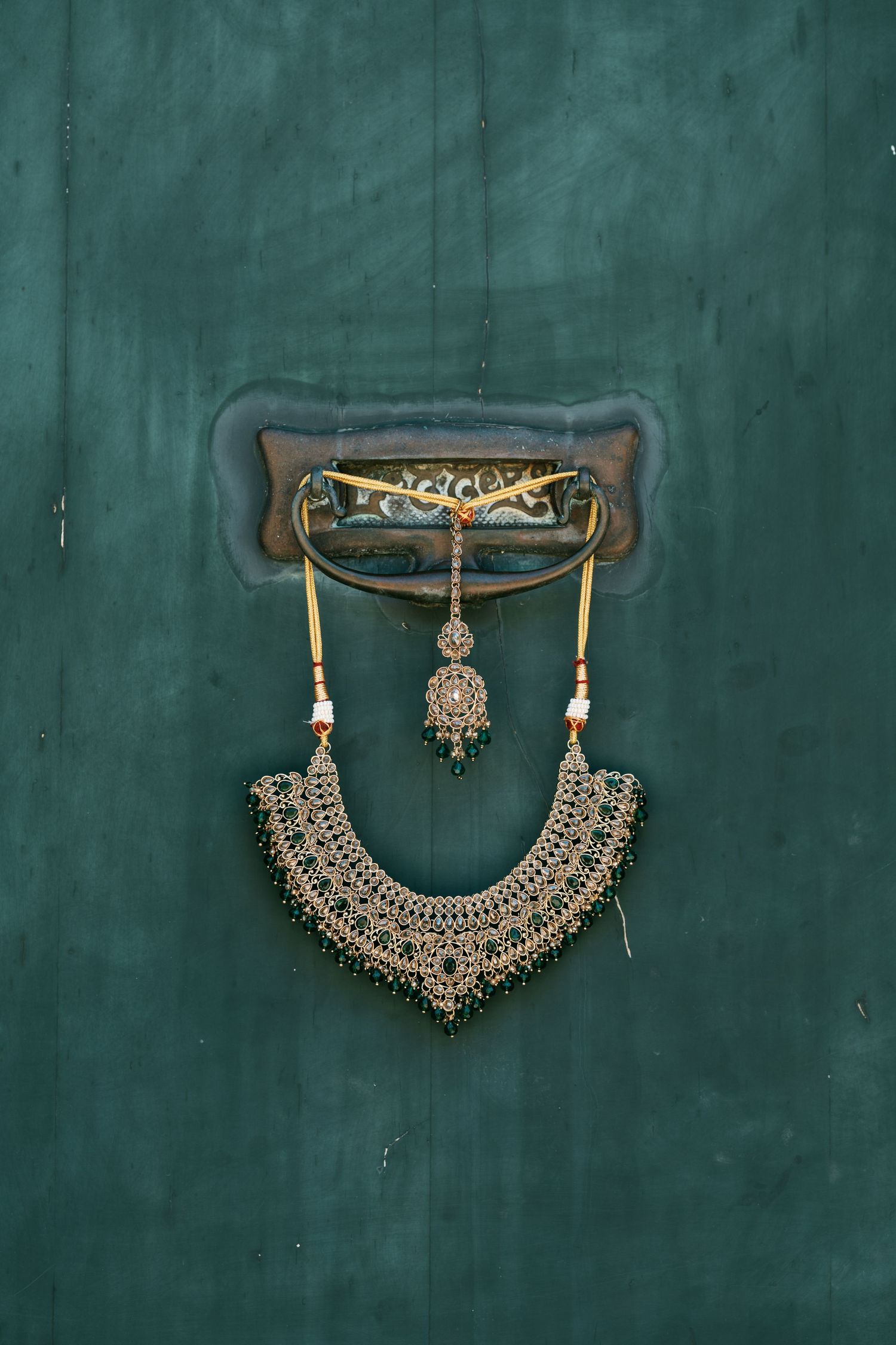 Ornate beaded bridal necklace with a tassel pendant hanging from a vintage brass door knocker on a deep green wooden door.