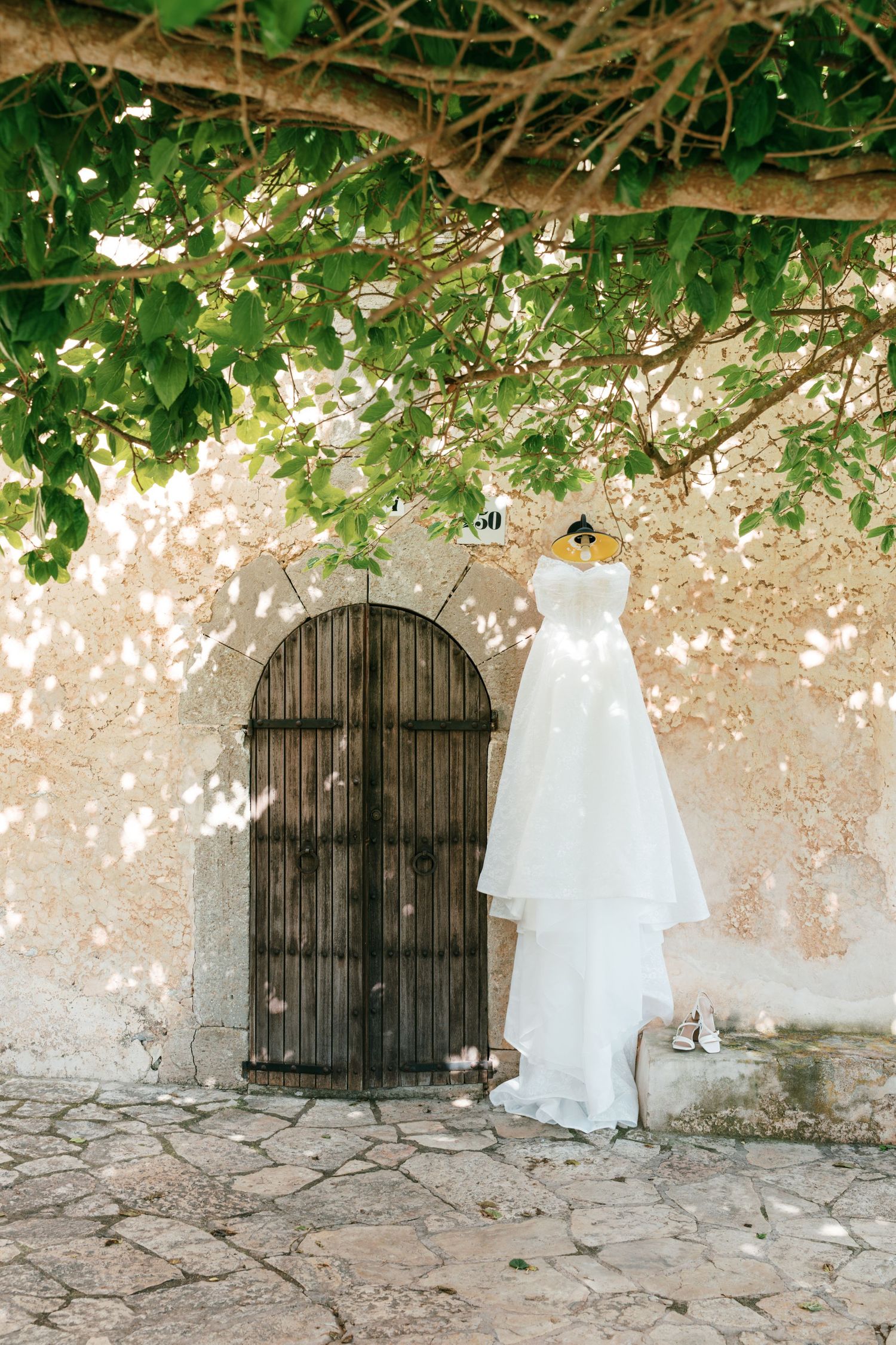 White wedding dress hanging on rustic wooden arched door beneath green tree, Mediterranean stone wall backdrop.