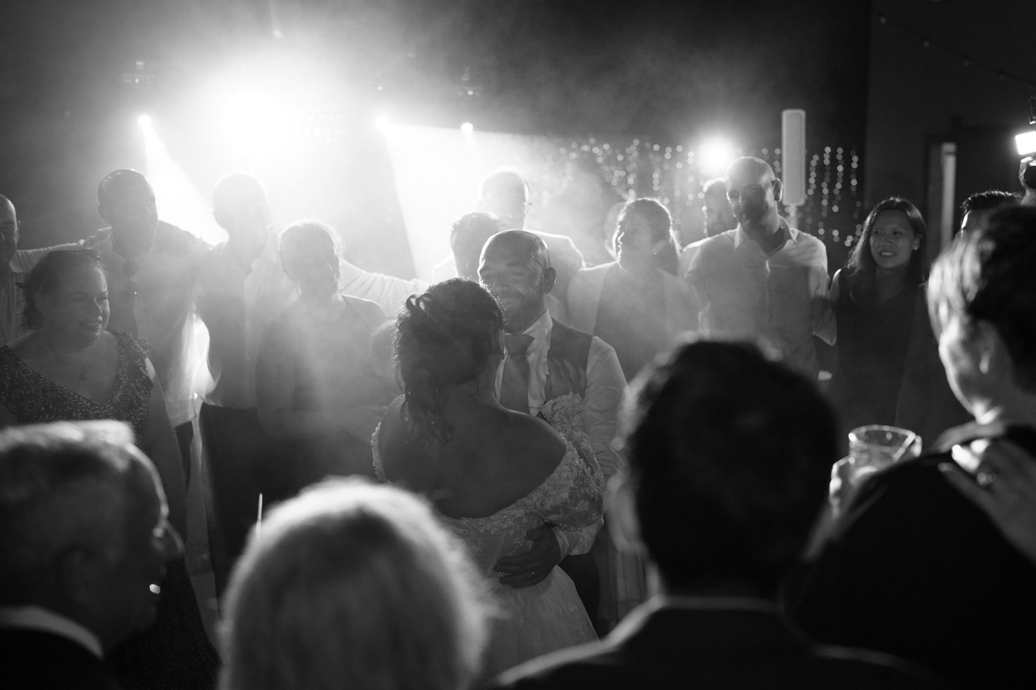 Black and white photo of a bride and groom dancing close as backlit wedding guests gather around them in a glowing, atmospheric light.