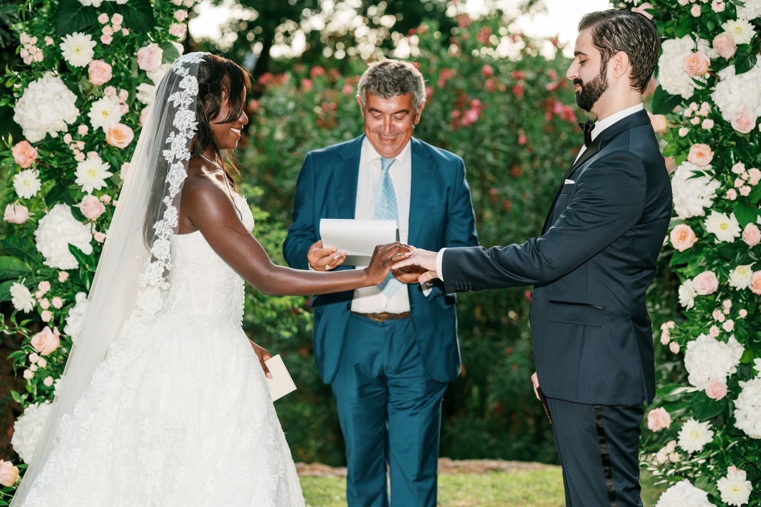 Bride in a lace veil and groom in a dark suit hold hands while an officiant reads vows during an outdoor garden wedding ceremony.
