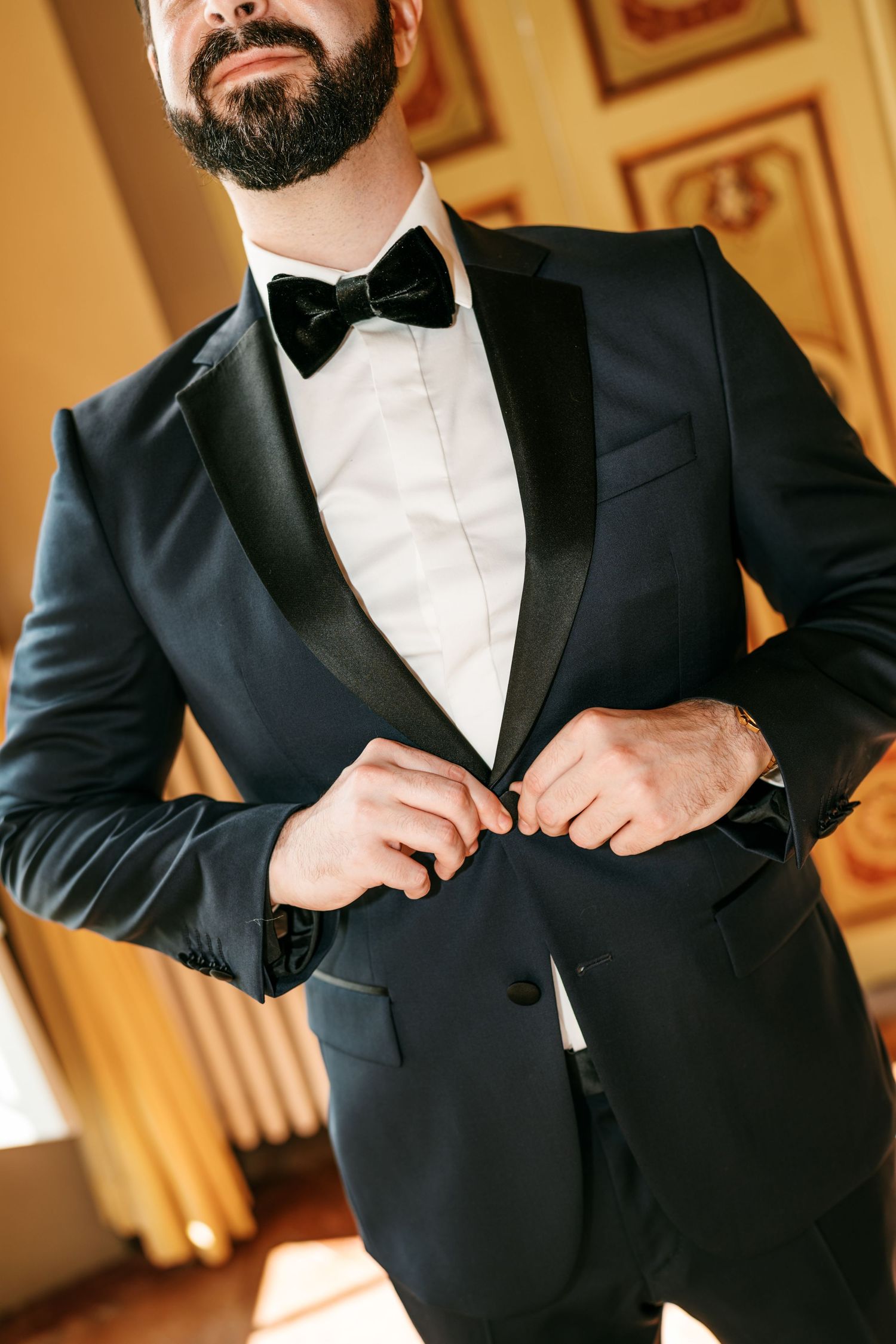 Bearded groom in a black tuxedo with satin lapels and a bow tie buttons his jacket in an ornate room with gilded frames.