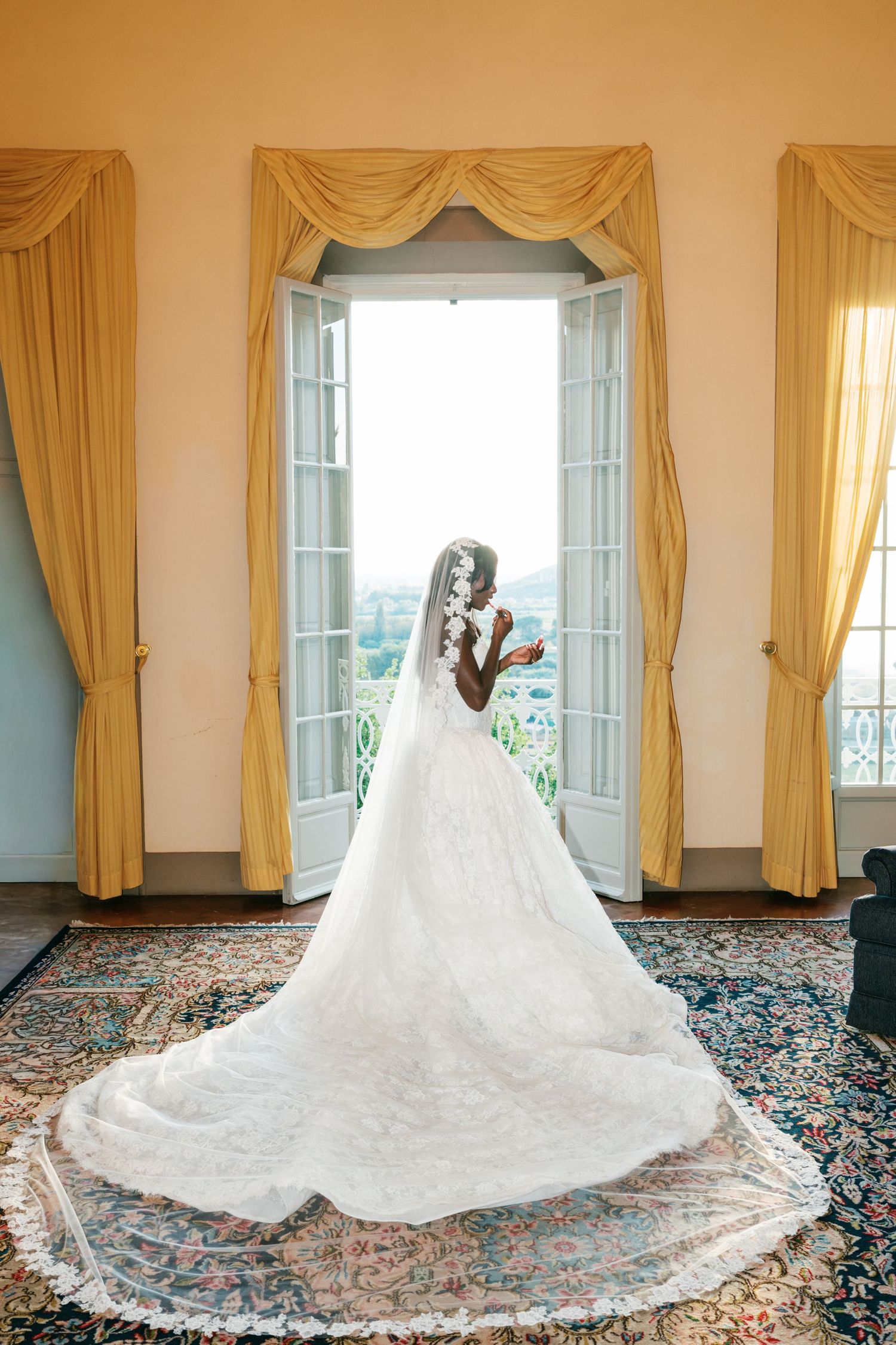 Bride in a lace wedding gown with a long cathedral veil stands at open French doors with golden drapes overlooking a scenic hillside.