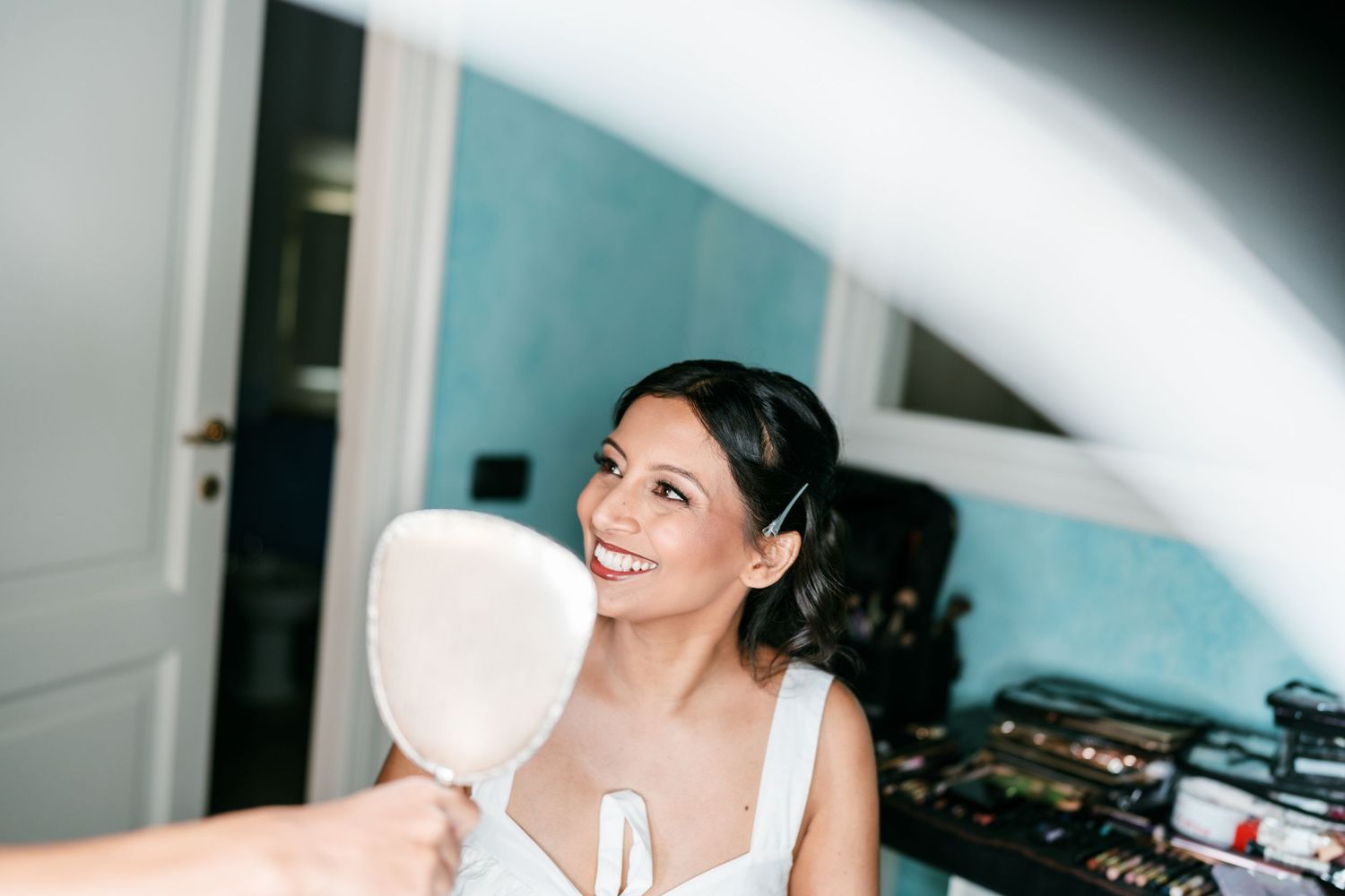 Smiling bride in a white dress holds a hand mirror while getting ready in a bright room with teal walls and makeup supplies nearby.