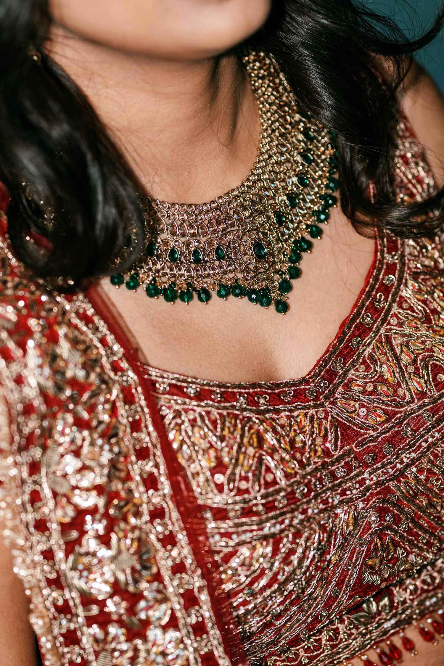 Close-up of a bride wearing an ornate red and gold embroidered lehenga blouse with a layered emerald bead and gold necklace.