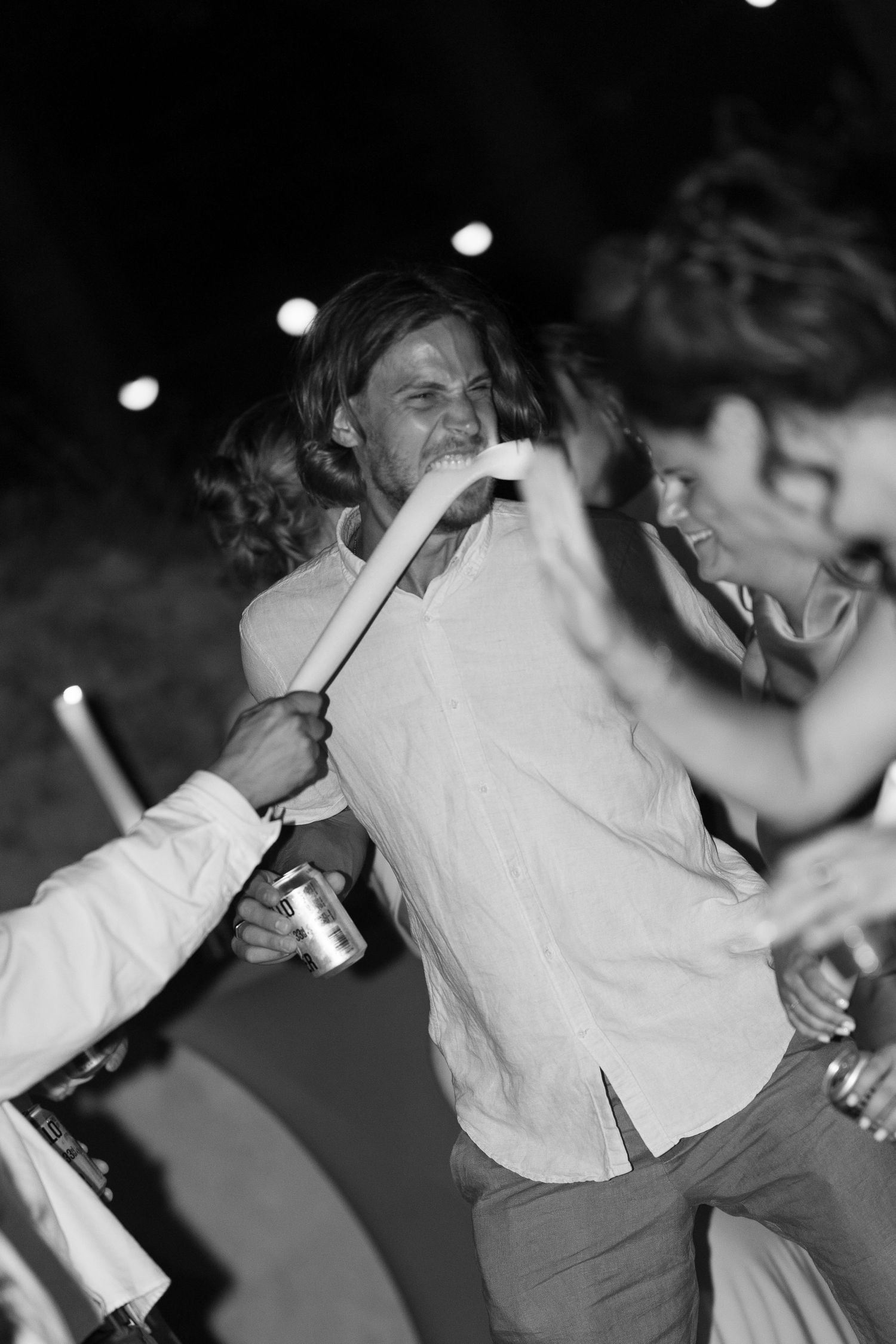 Black and white photo of long-haired man drinking from a tube at a lively outdoor party.