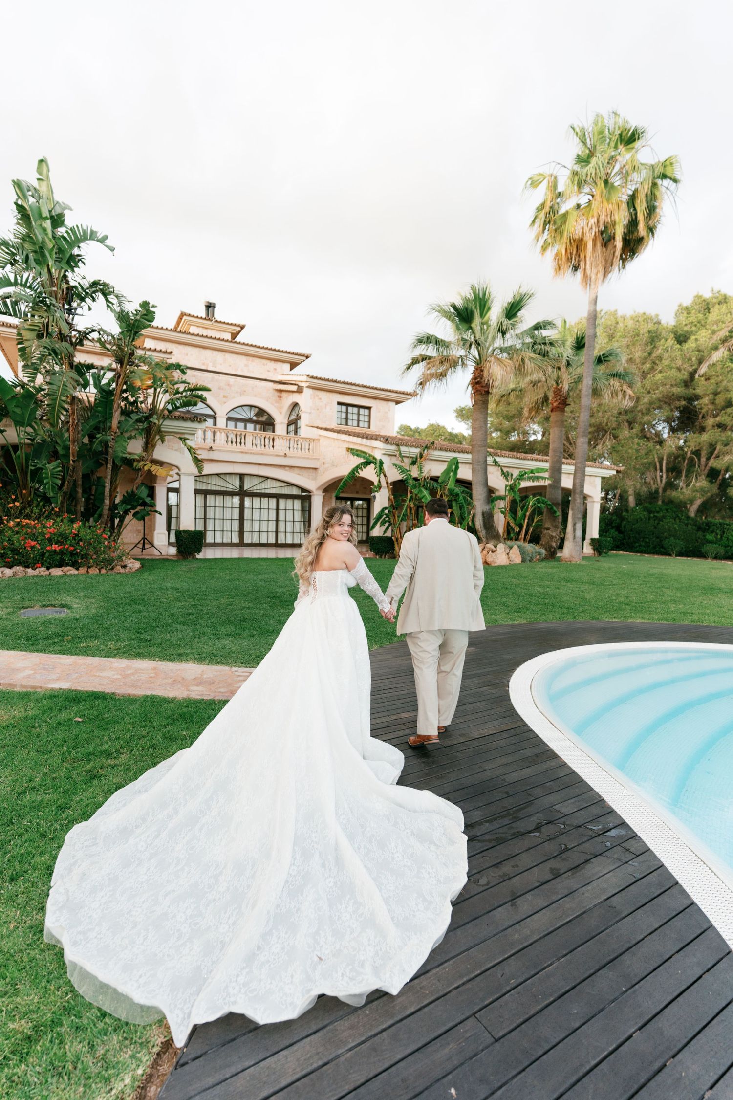 Bride in flowing lace gown walks hand-in-hand with groom past pool at luxury villa wedding venue.