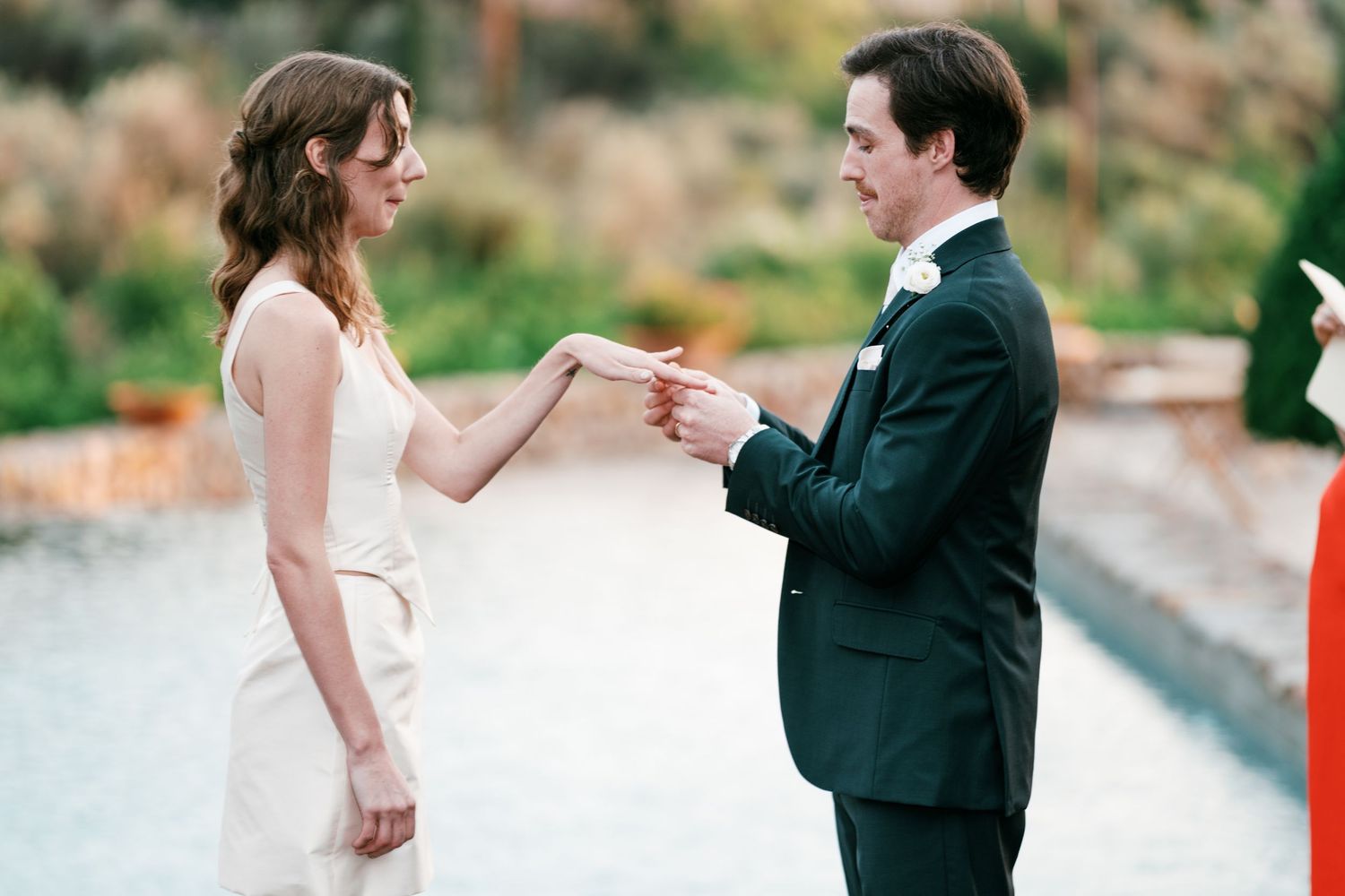 A bride in a white dress and a groom in a dark suit exchange rings during an outdoor wedding ceremony beside a pool.