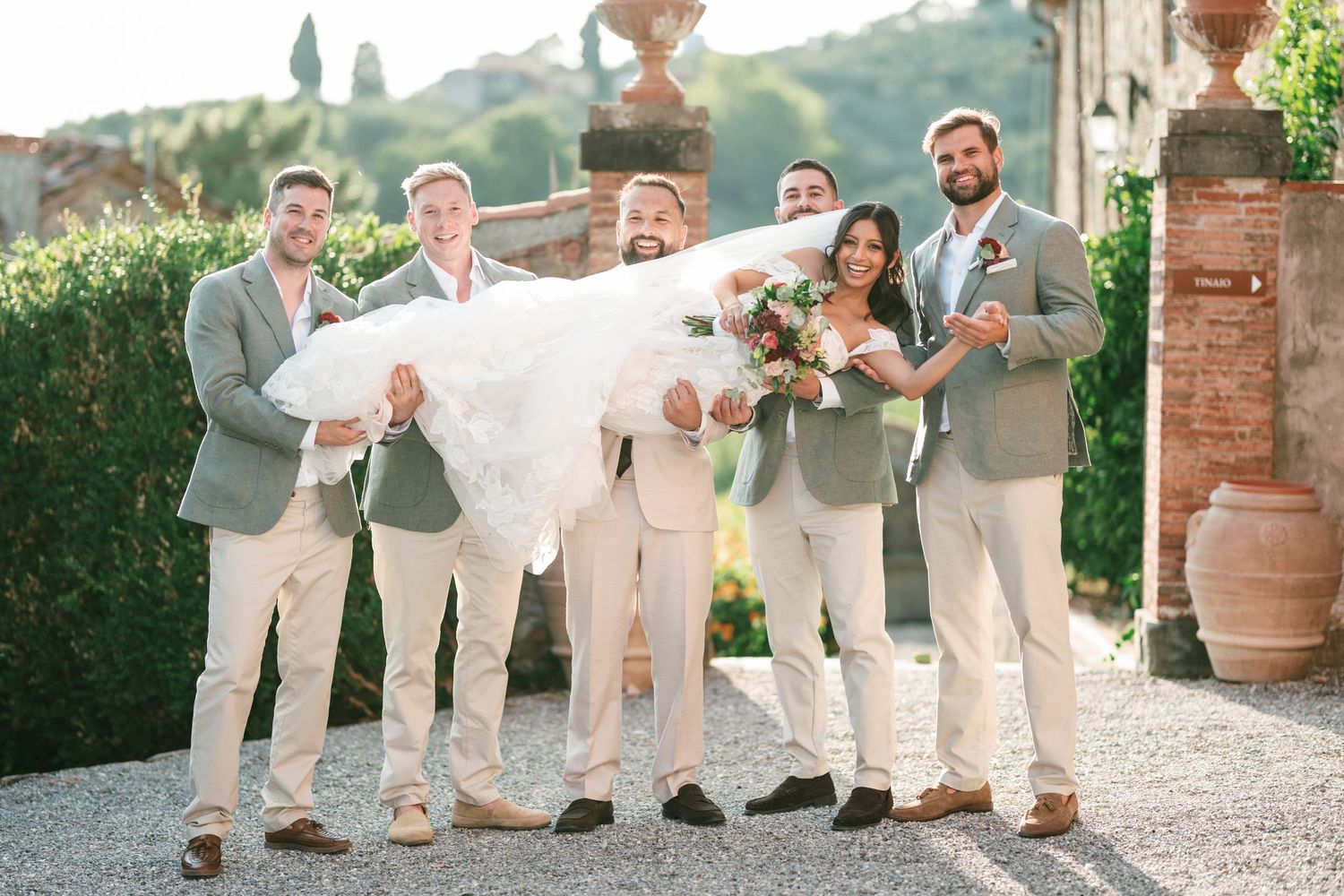 Groomsmen in tan suits playfully lift a bride in a white veil and bouquet at a sunny Tuscan villa wedding with terracotta pots and greenery.