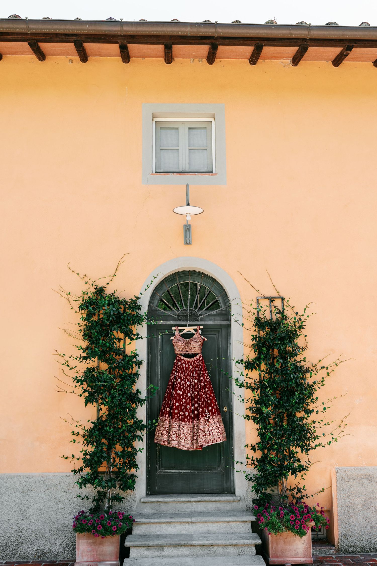 A red embroidered lehenga hangs on a hanger from the arched green door of a peach Tuscan villa, framed by climbing vines and flower pots.