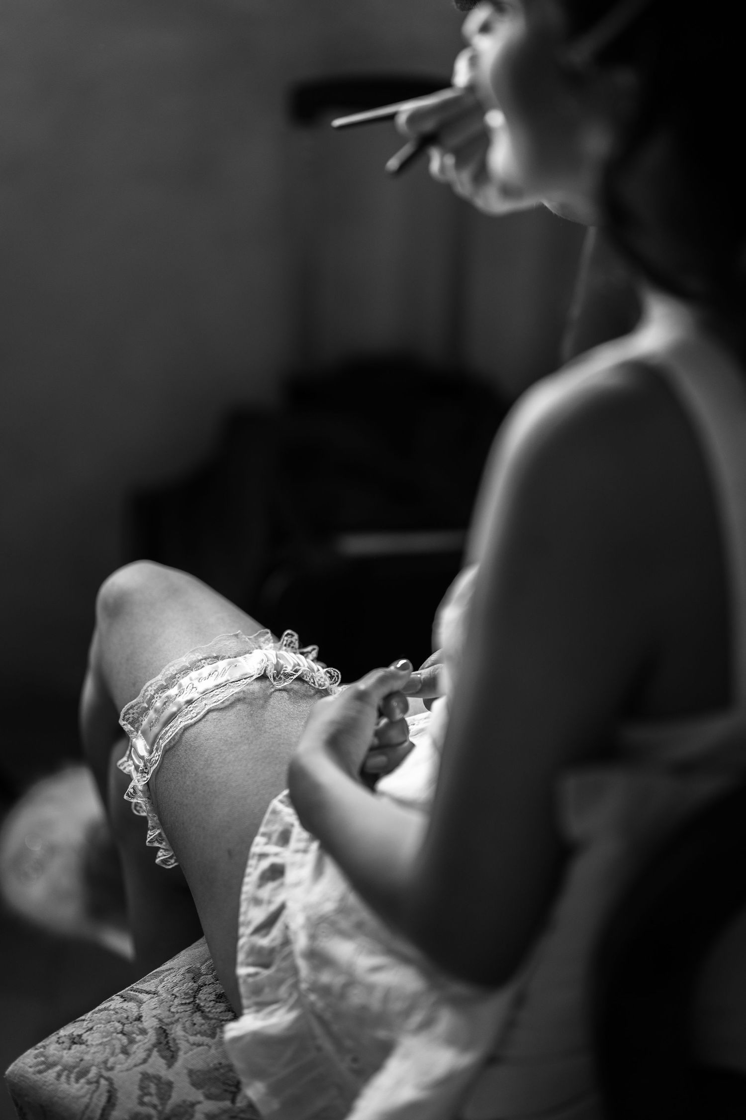 Black and white photo of a bride seated in soft light, adjusting a lace wedding garter on her thigh while getting ready.
