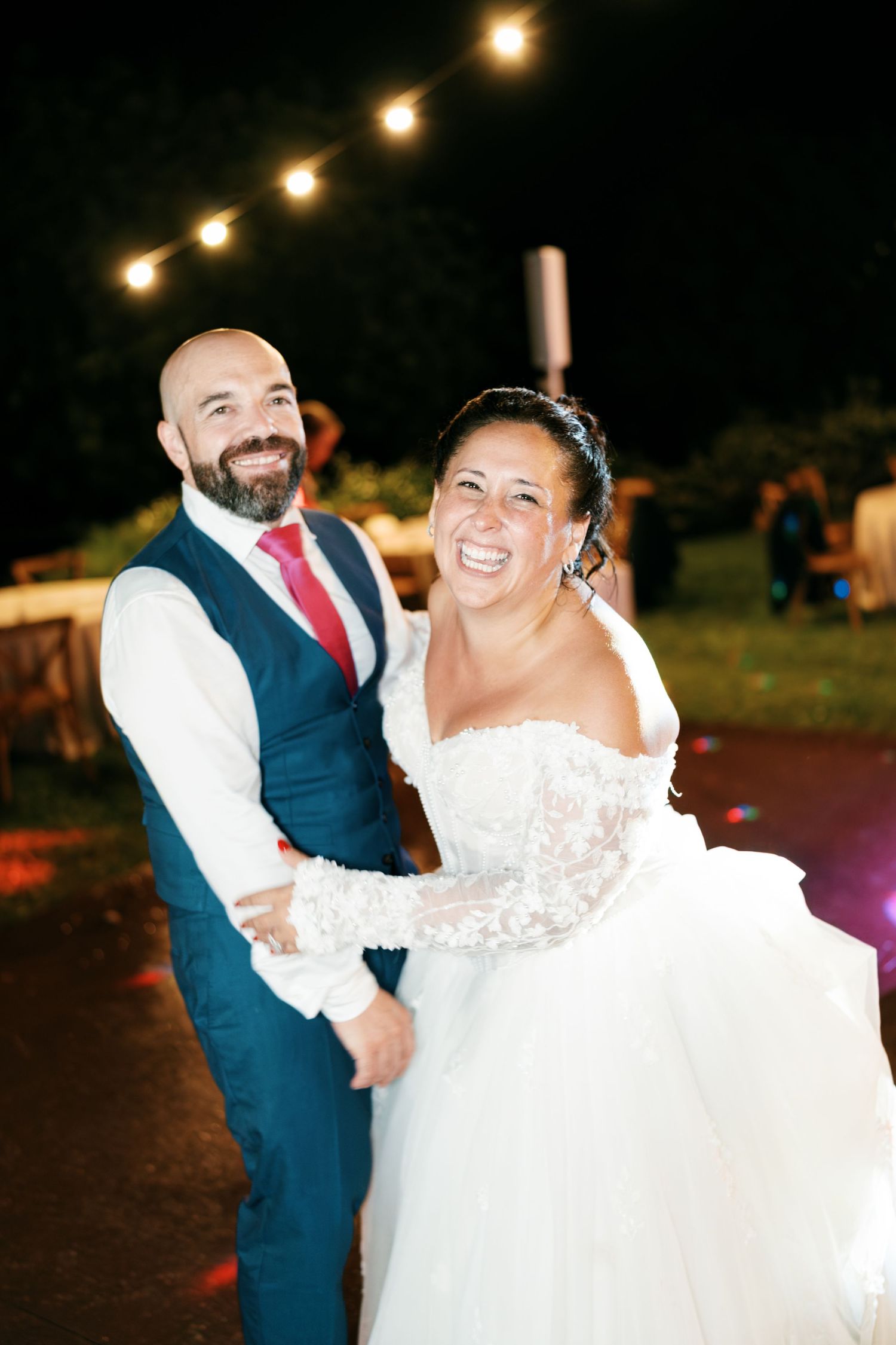 Smiling bride in a lace off-shoulder gown stands beside a bearded groom in a navy vest and red tie at a night wedding under string lights.