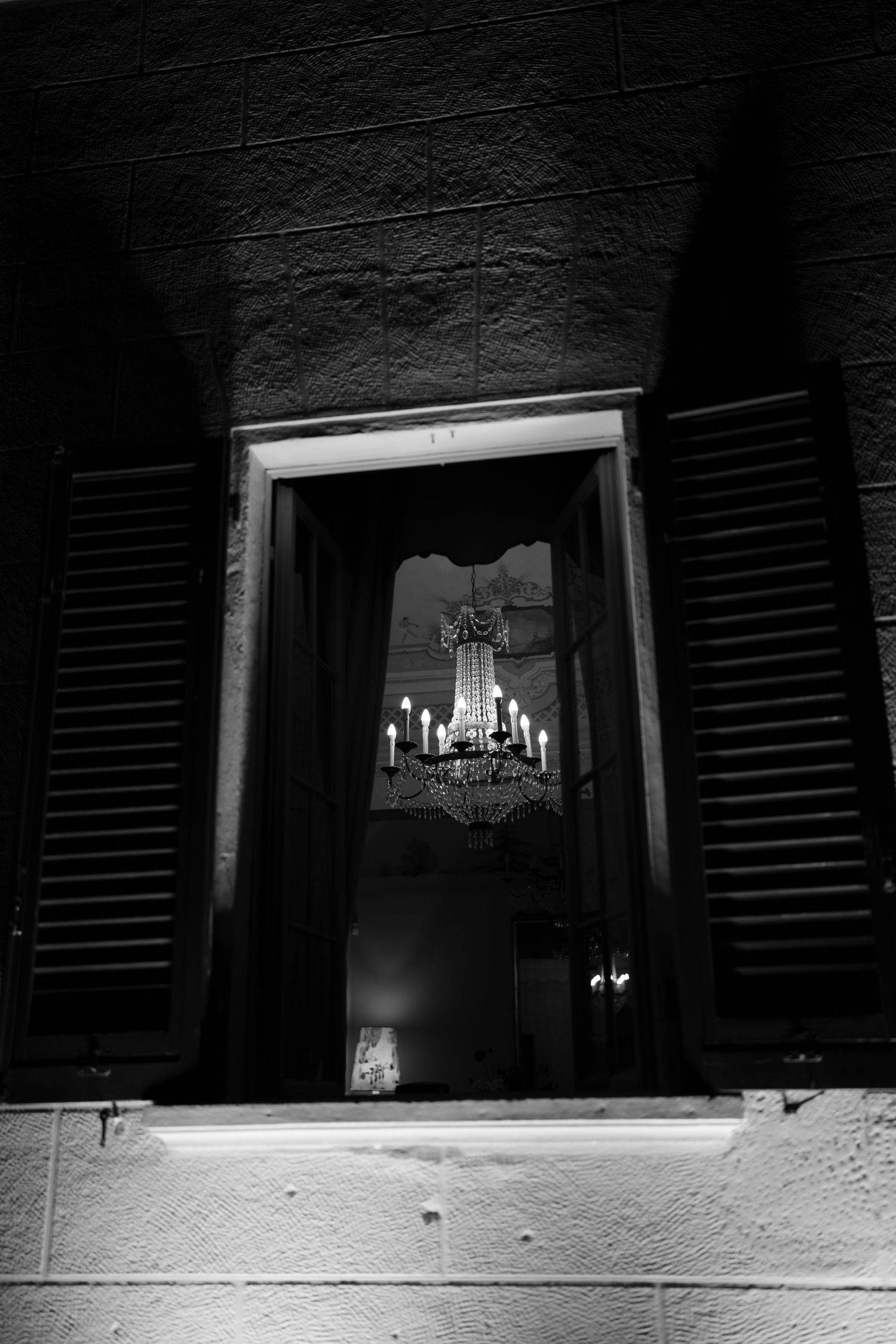 Black and white view through an open window with wooden shutters revealing a glowing crystal chandelier inside.