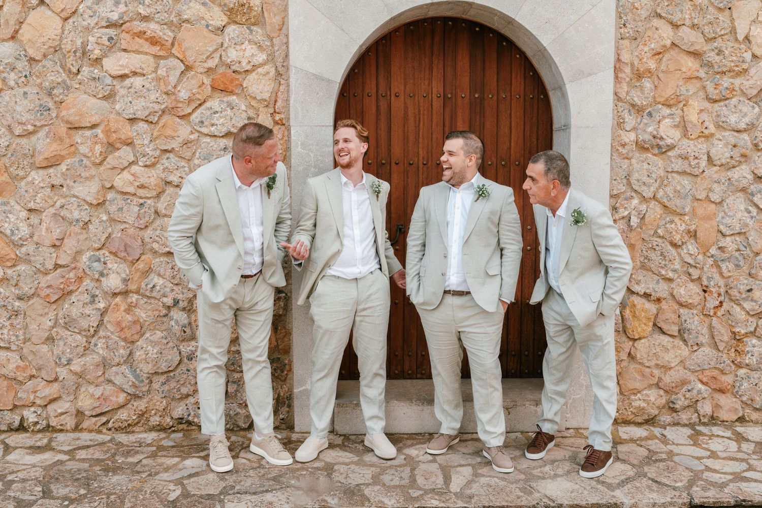 Four groomsmen in matching beige linen suits pose and laugh together outside a rustic stone chapel. Mallorca wedding photographer