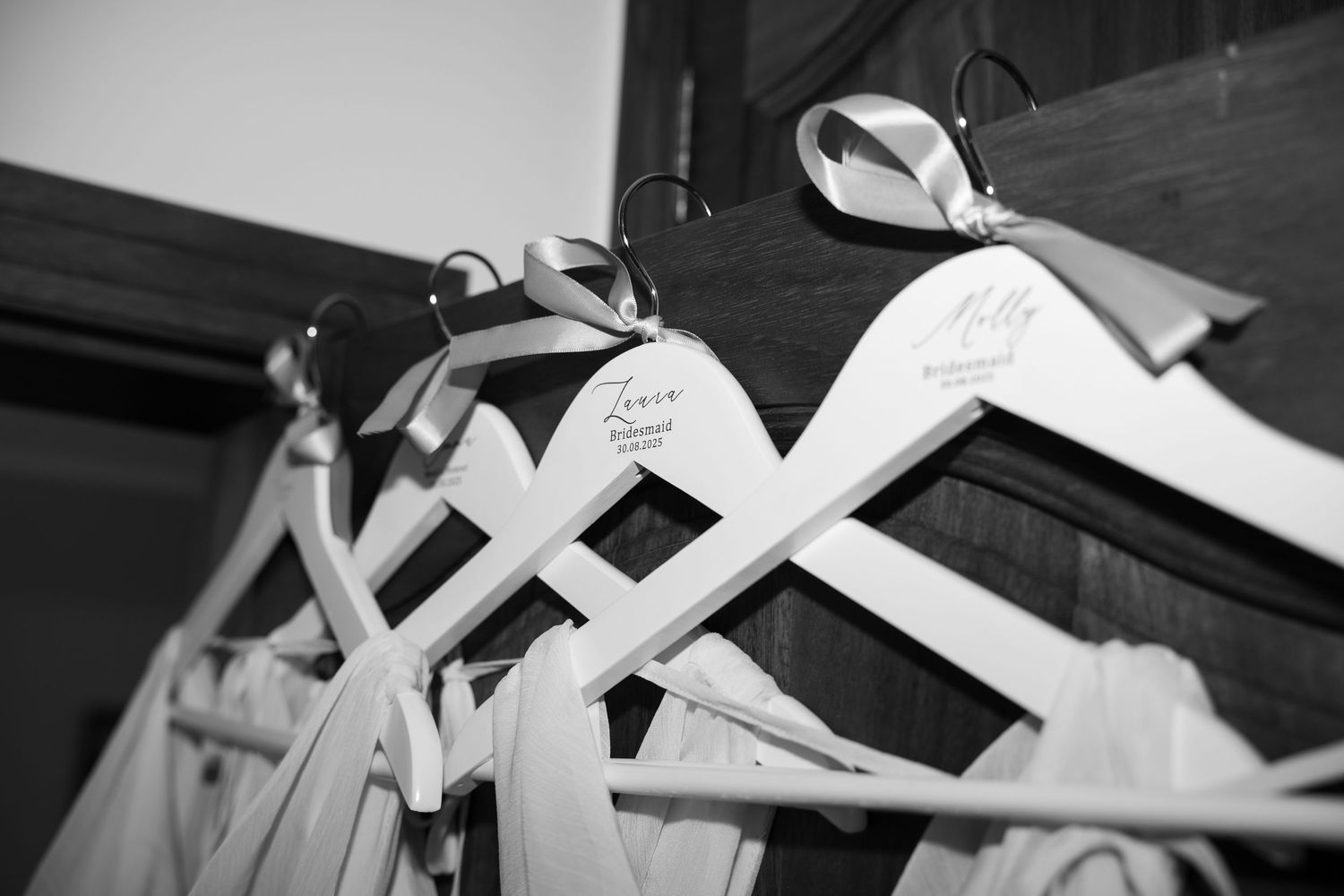 Black and white photo of personalized white wedding dress hangers with ribbon bows hanging on a rustic wooden rack. Mallorca wedding film