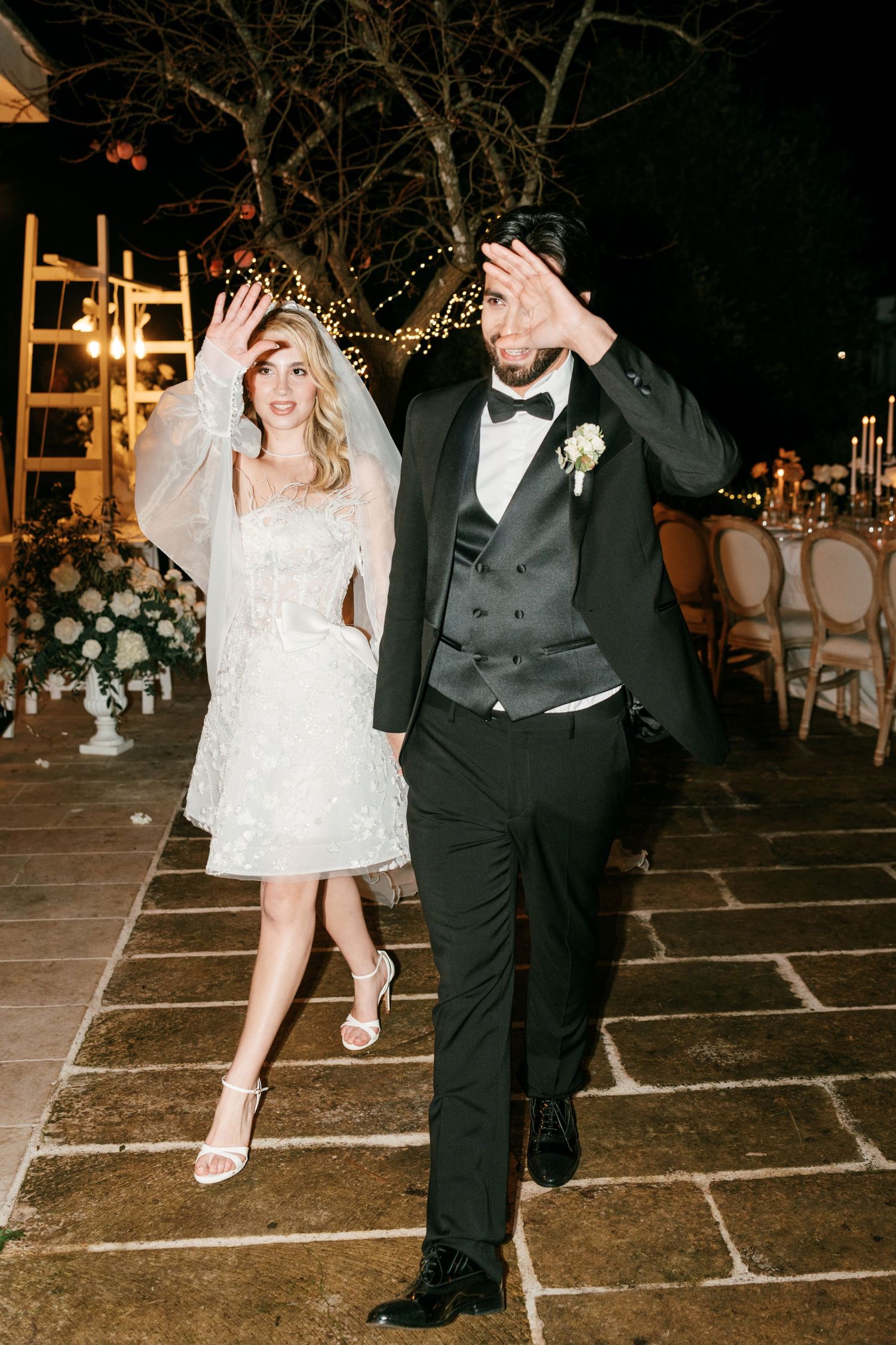 Bride in short lace dress and groom in black tuxedo make grand entrance at outdoor evening wedding reception. Puglia wedding photographer,
