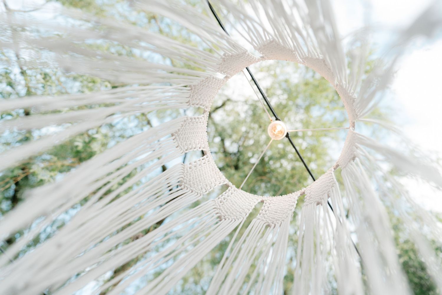 Elegant white macrame wedding decoration hanging outdoors with string lights against a canopy of green trees. Mallorca wedding photographer