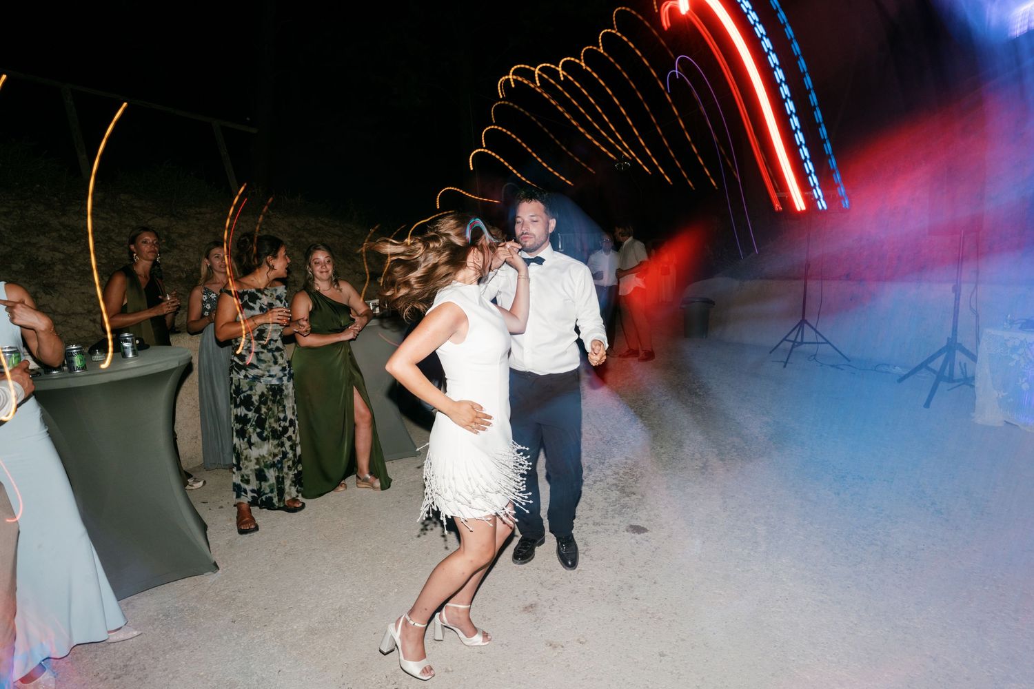 Bride and groom share first dance outdoors at night with colorful lights and smoke effects at wedding reception.