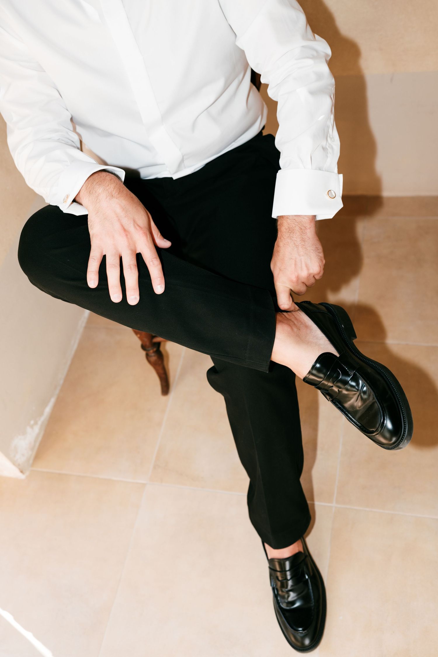 Man in white dress shirt and black trousers sitting cross-legged wearing polished black leather loafers.