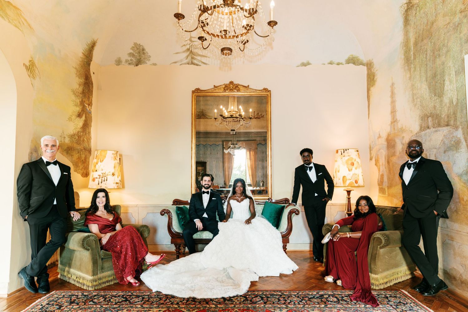 Bride in a white gown poses with groom, groomsmen and bridesmaids in red dresses in an ornate Tuscan villa room.