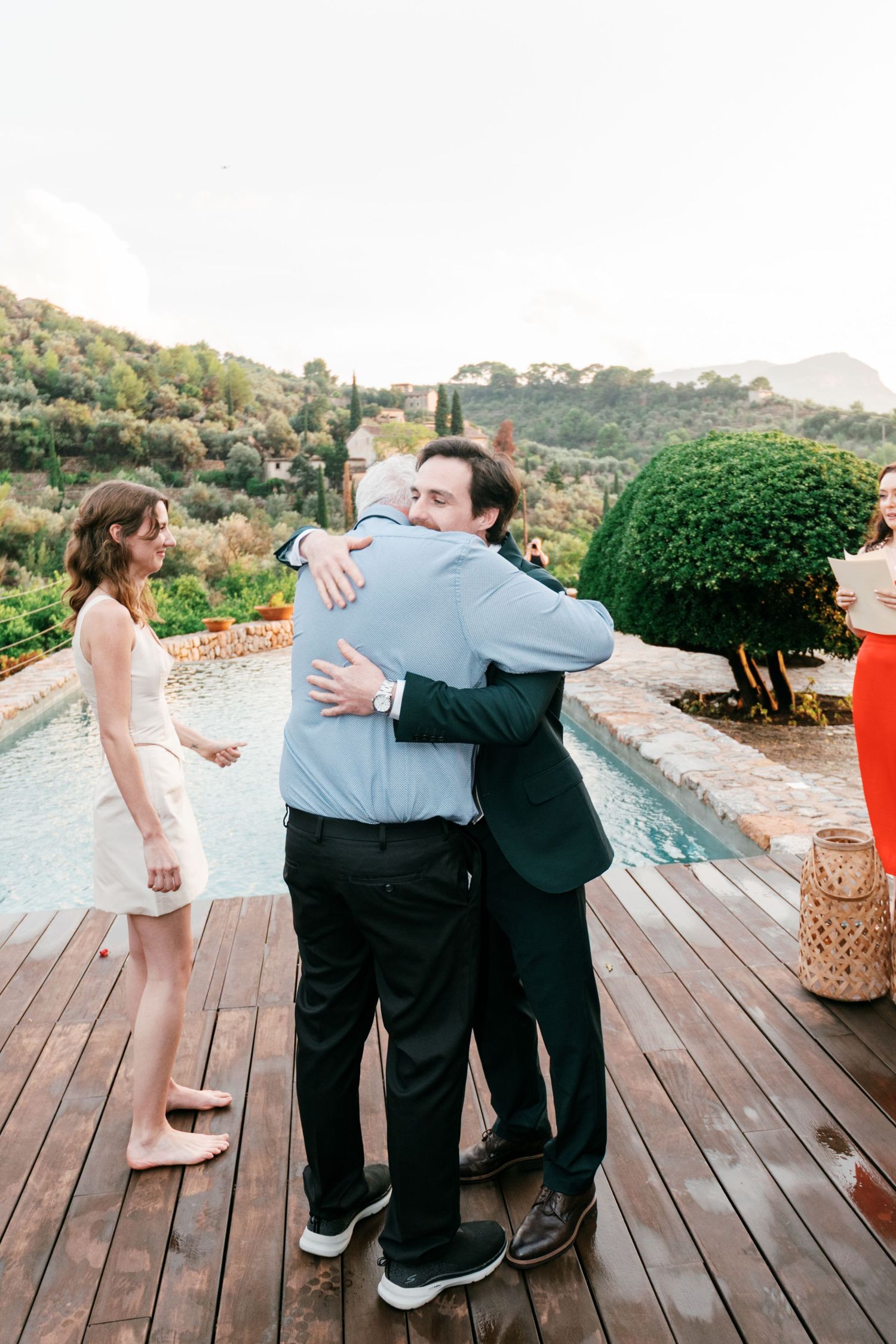 Two men embrace on a wooden pool deck at an outdoor wedding as a barefoot woman in a cream dress looks on, with green hills behind.