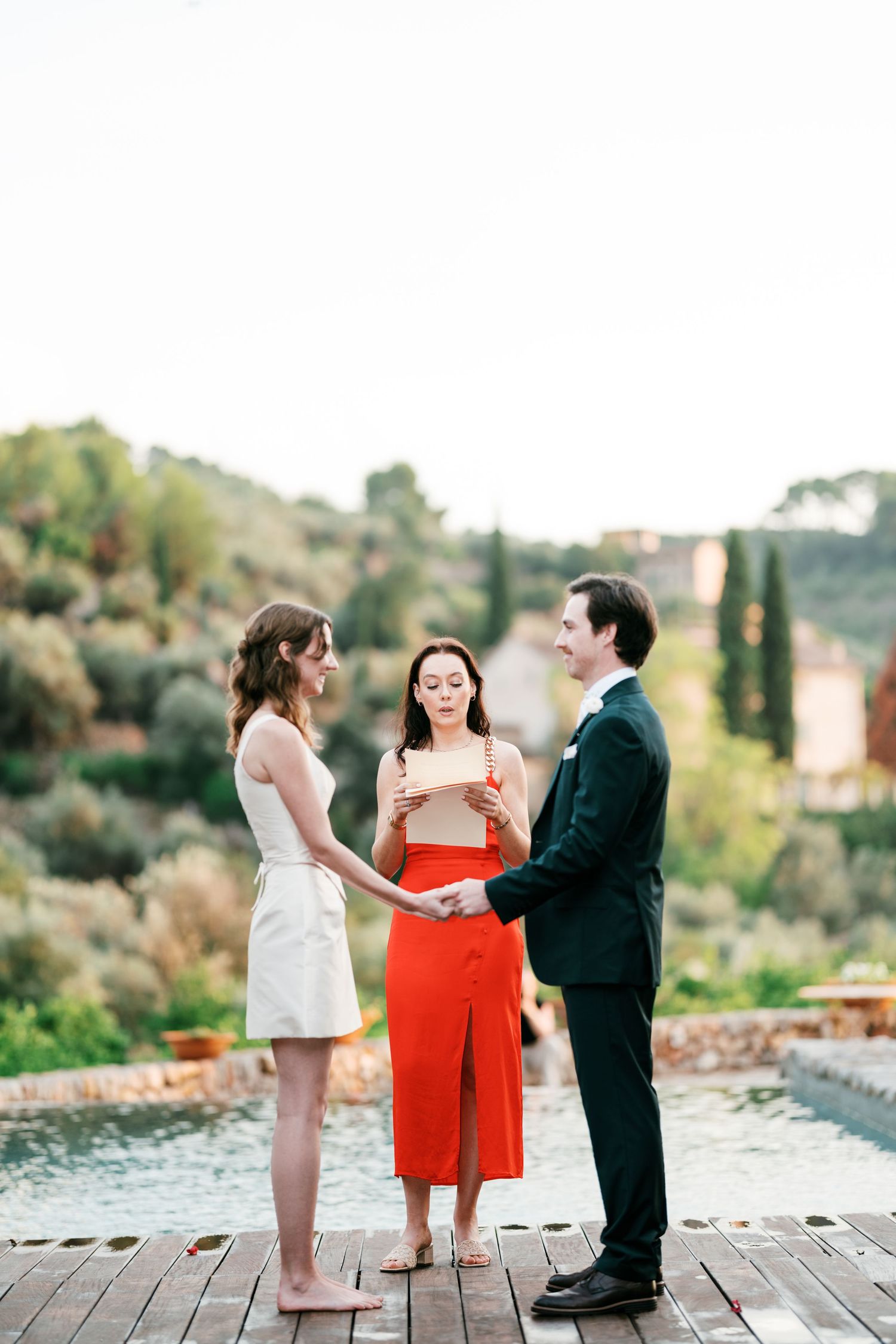 Bride and groom hold hands outdoors while an officiant in a red dress reads vows beside a pool with scenic hillside views.