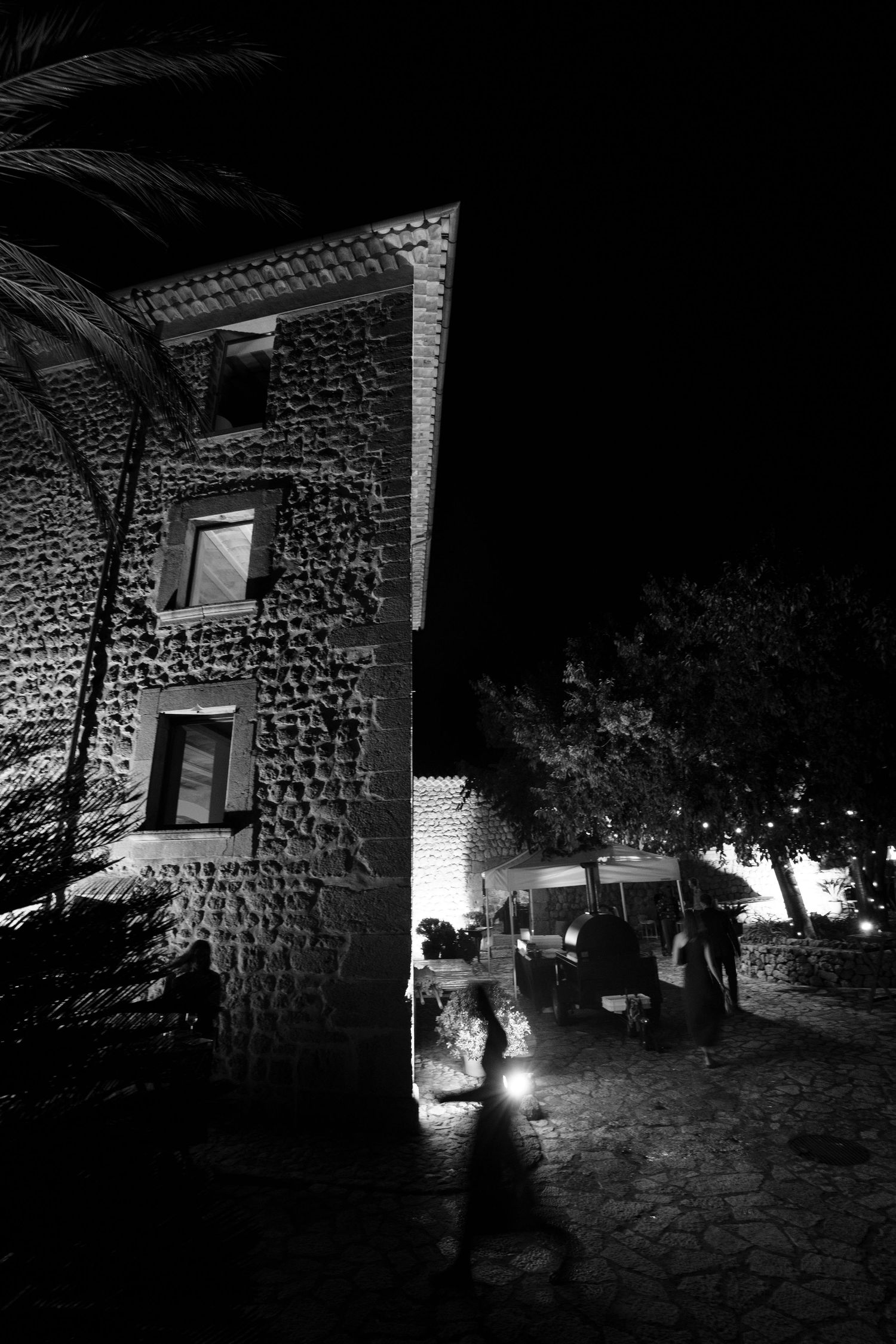 Black and white night photo of a rustic stone building with lit windows, palm fronds and people gathered in a cobblestone courtyard.