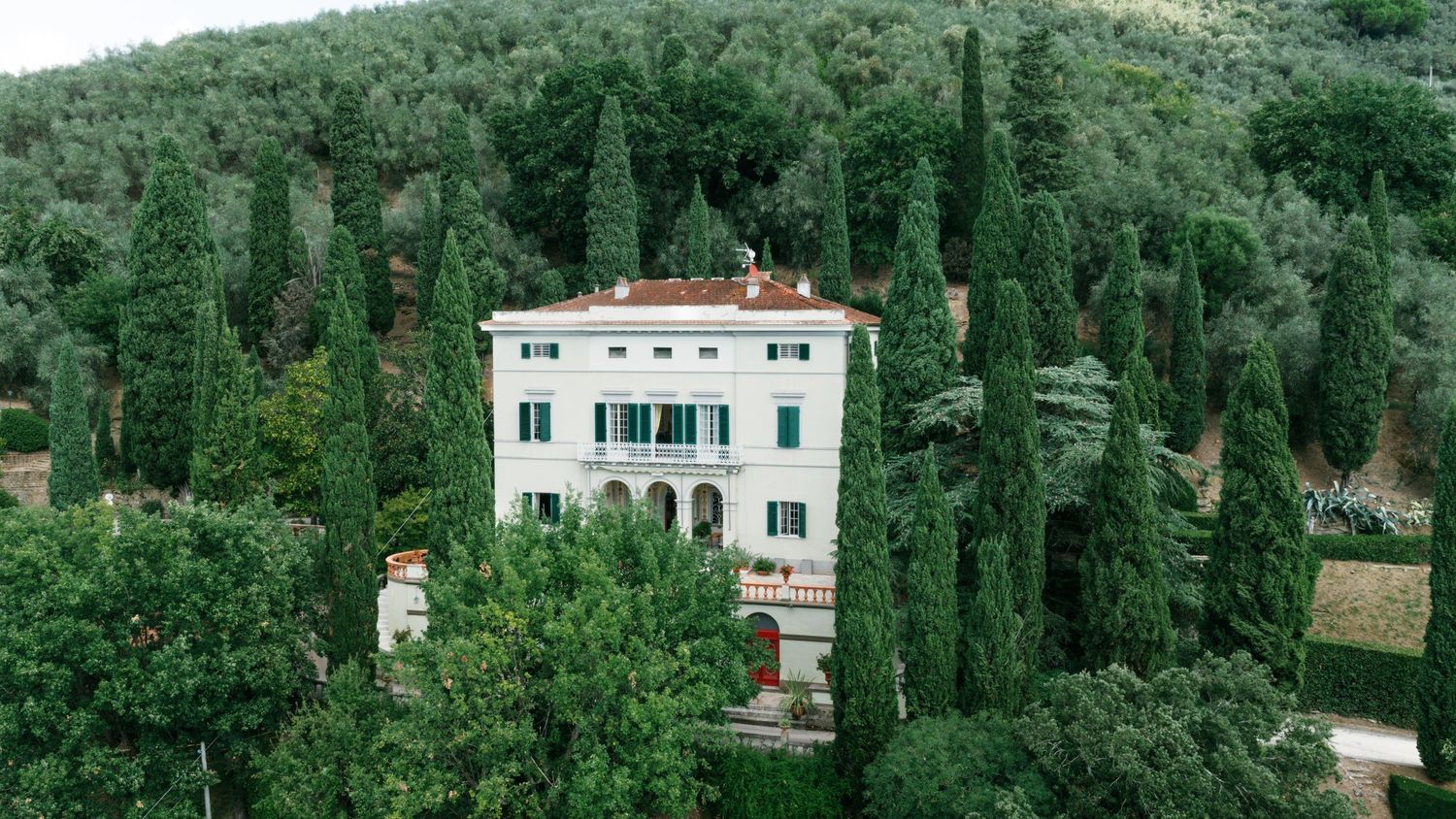 Aerial view of an elegant Tuscan villa with a terracotta roof surrounded by tall cypress trees on a lush green hillside.