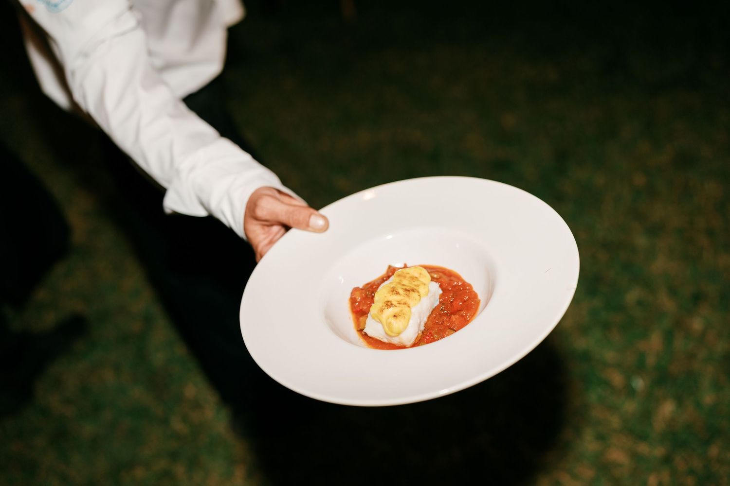 Waiter in a white shirt presents a wide white bowl of tomato sauce topped with a seared fish fillet at an outdoor evening event.