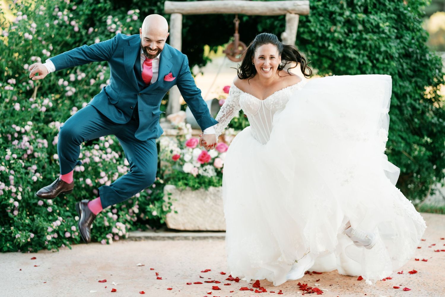Joyful bride in a white ball gown and groom in a blue suit leap through the air on a petal-strewn path beside a flowering garden.