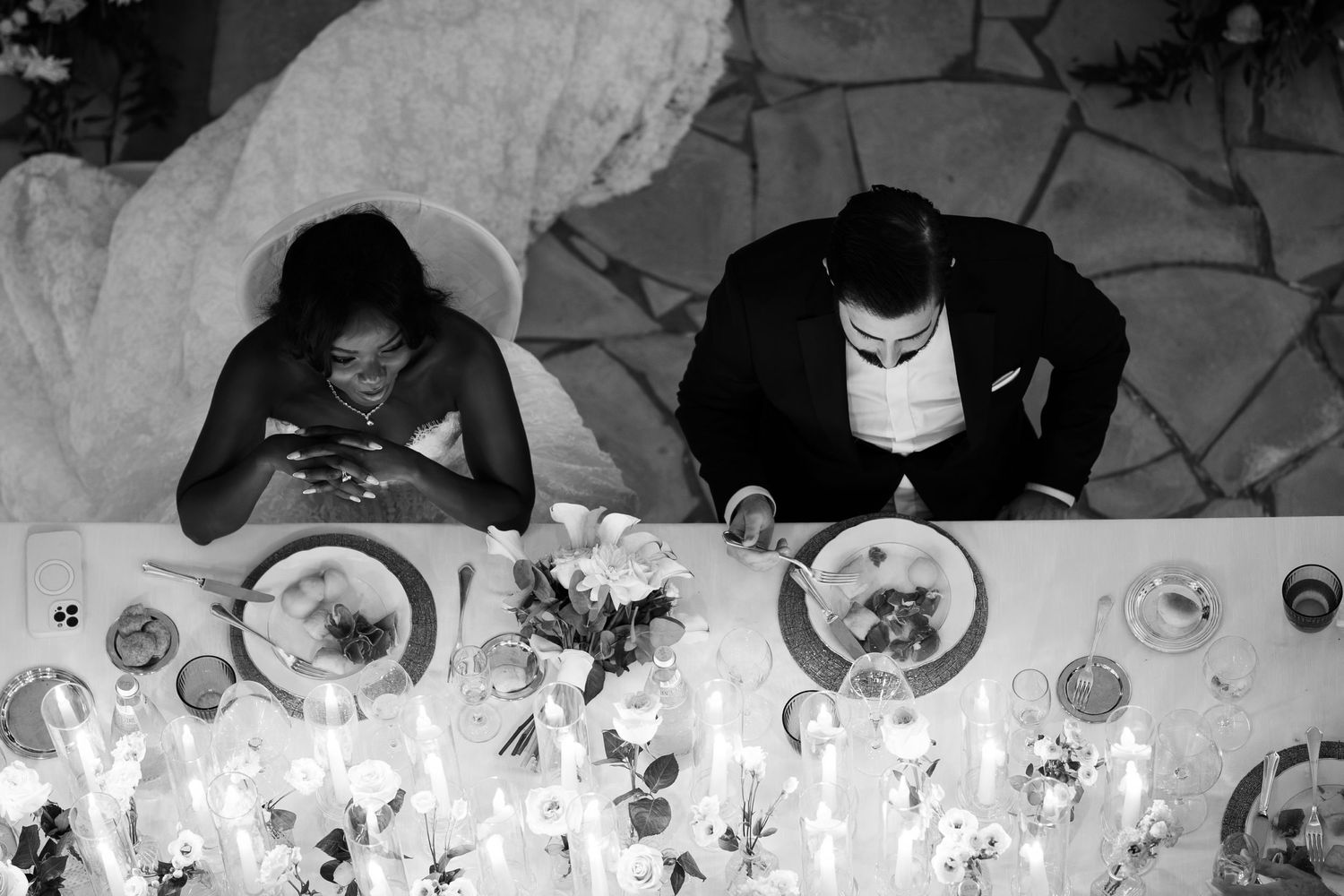 Black and white top-down shot of a bride and groom seated at a candlelit wedding table with plates, flowers and glassware.