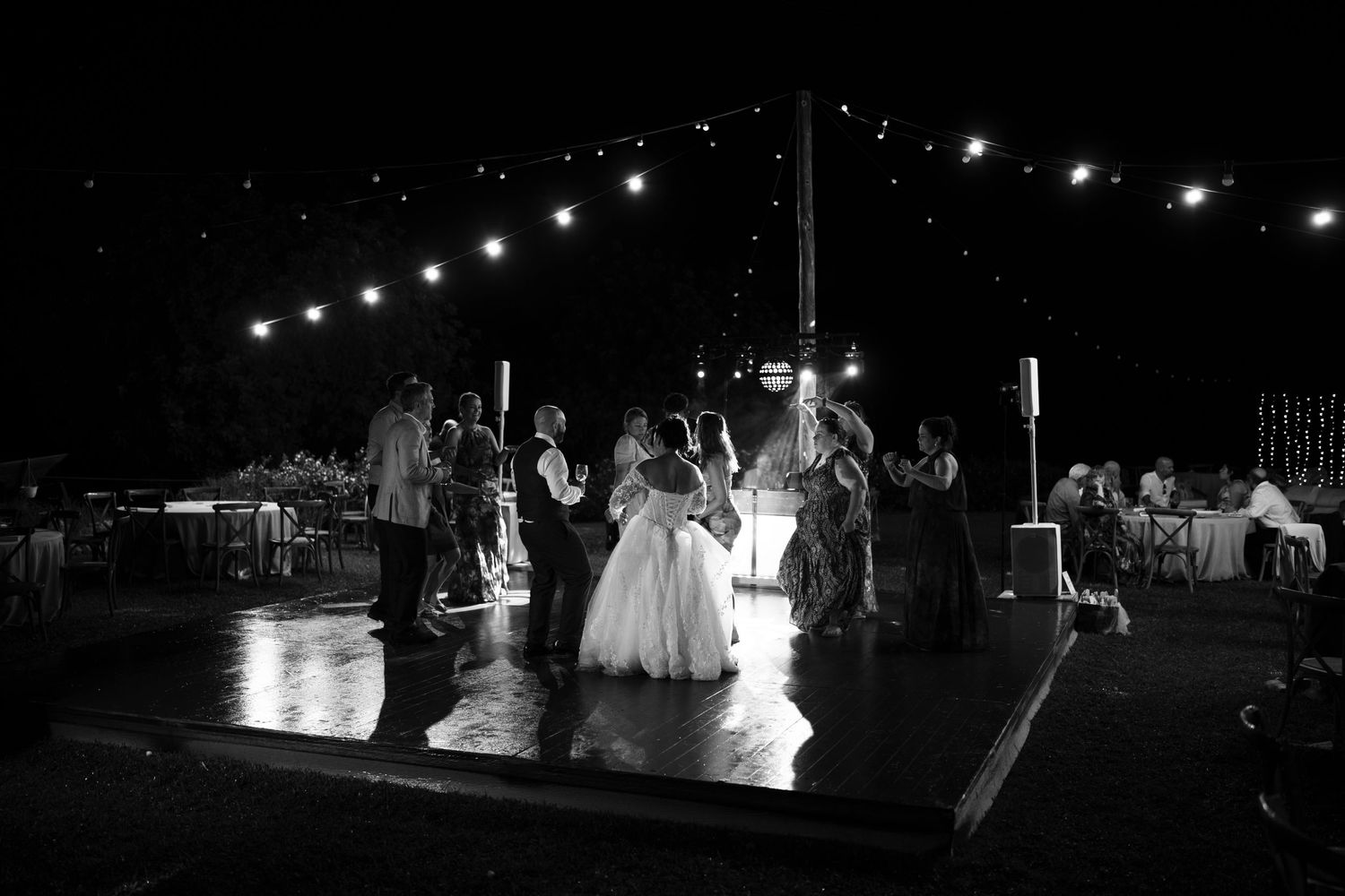 Wedding guests gather around the bride and groom on a dance floor at a night reception lit by string lights.