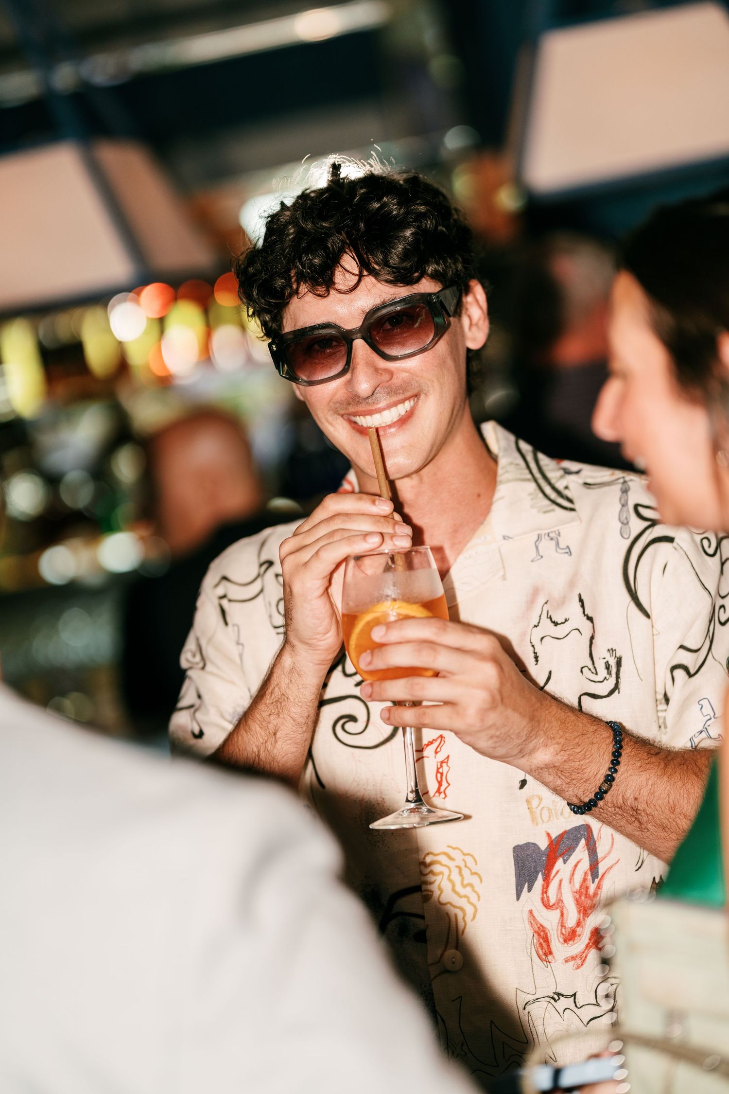 Smiling man in sunglasses and a patterned shirt sips an orange cocktail through a straw at a lively, warmly lit bar.