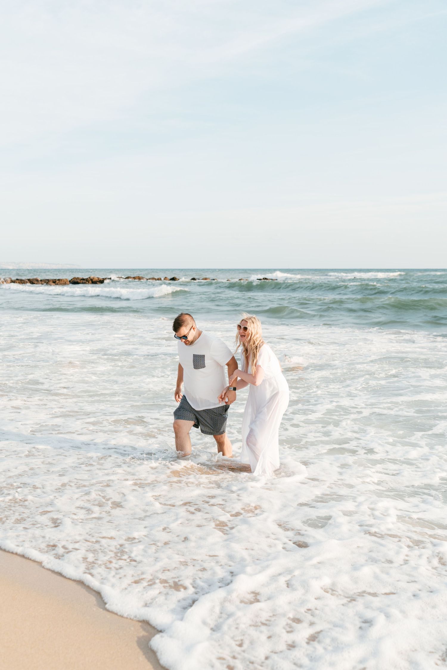 A couple holds hands and laughs while ocean waves wash over their feet on a sunny beach. Mallorca wedding filmmaker, videographer