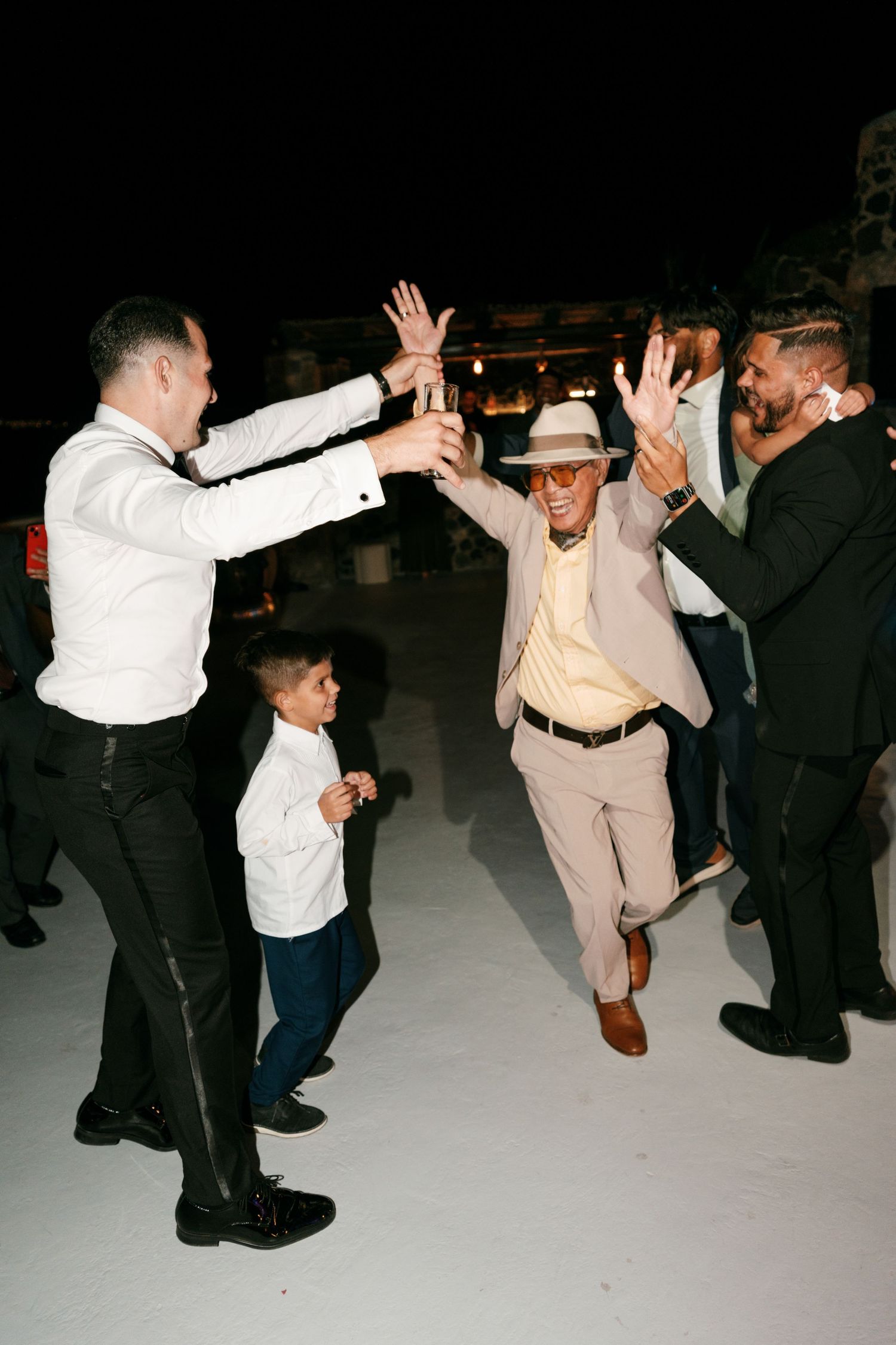 Men and a young boy dancing joyfully at an outdoor evening celebration, with one man in a tan suit and hat.