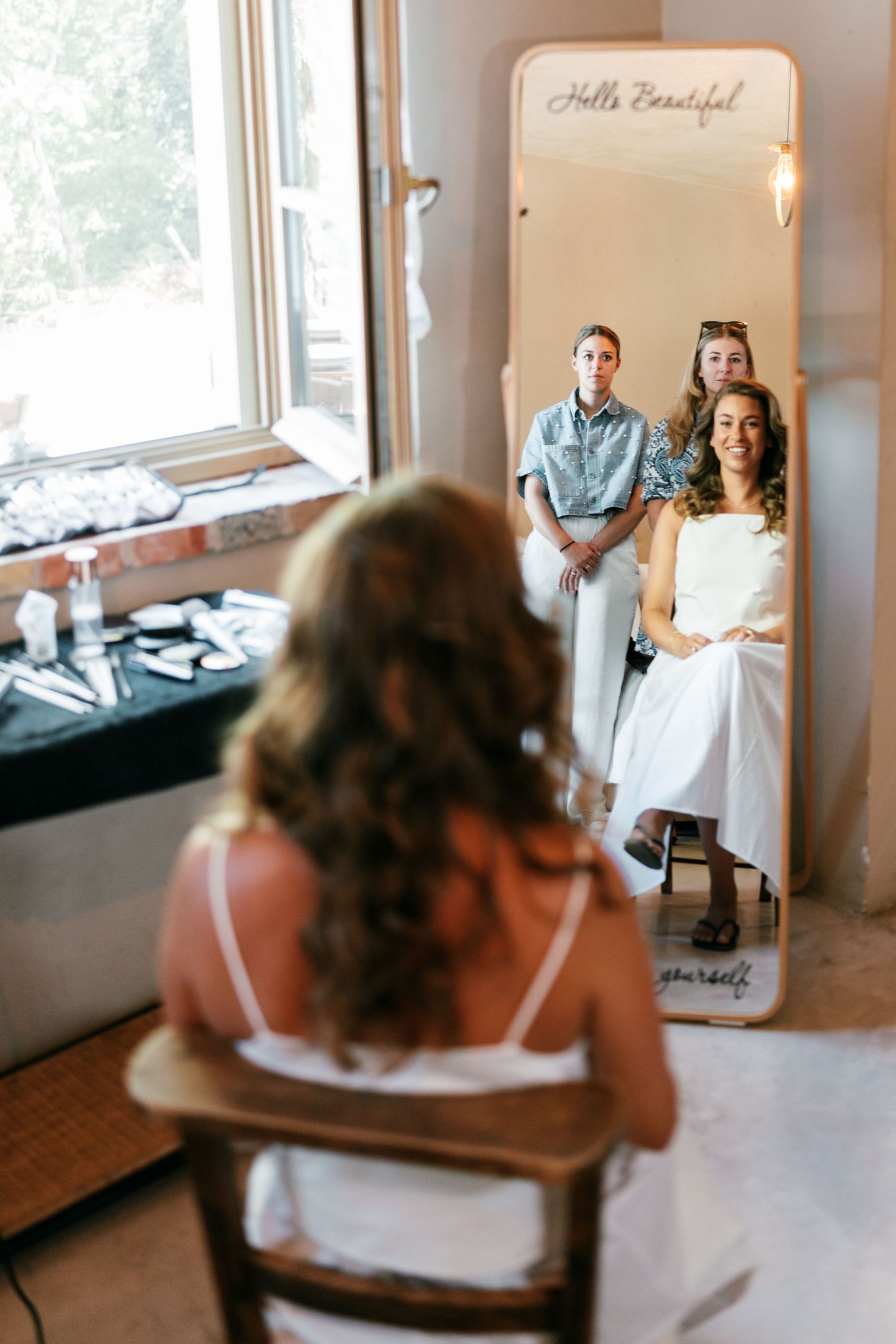 Bride sits in chair viewing mirror reflection during wedding preparations with bridesmaids in bright dressing room.