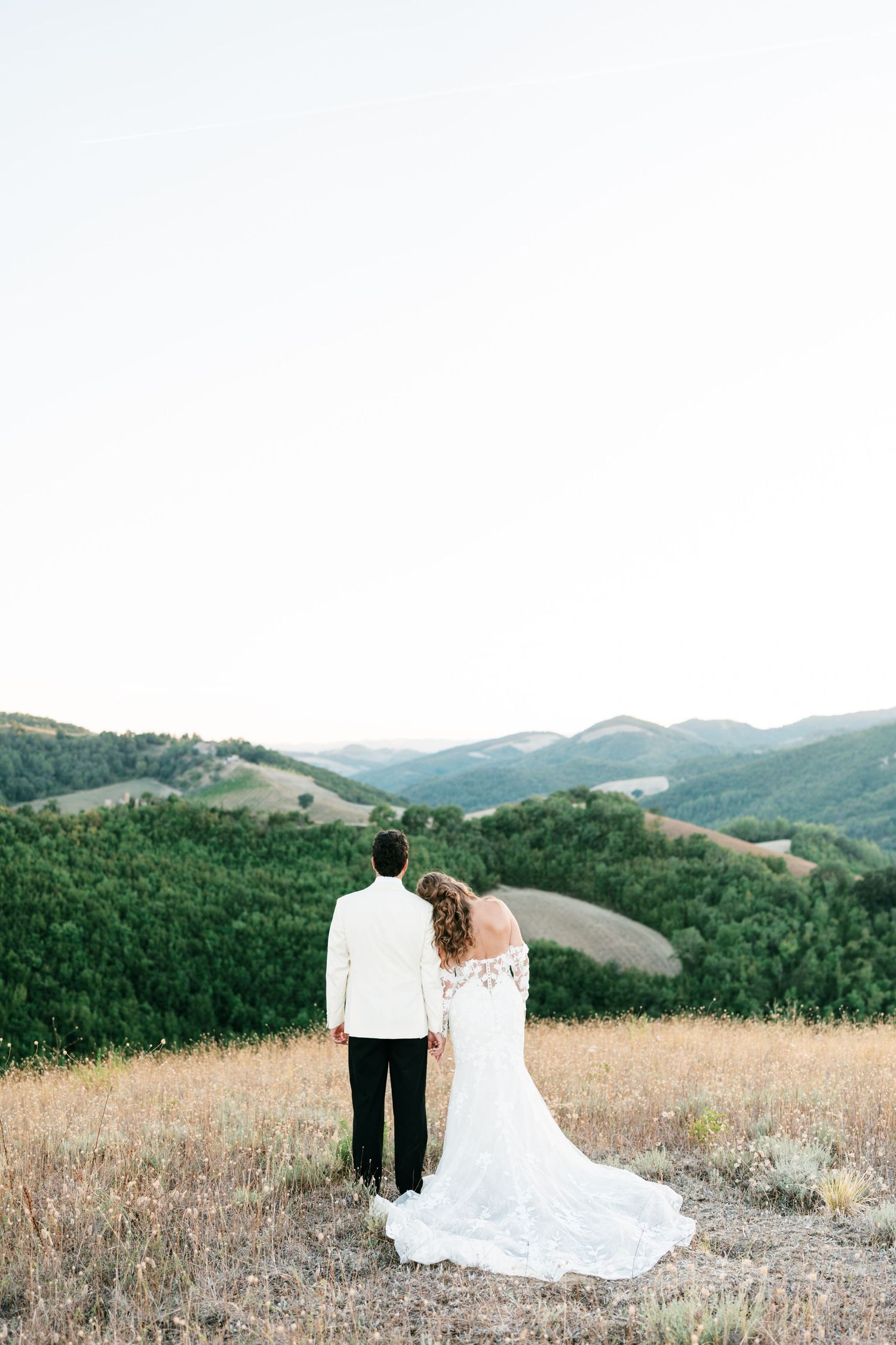 Bride and groom stand hand in hand overlooking lush green rolling hills at golden hour in a romantic outdoor wedding portrait.
