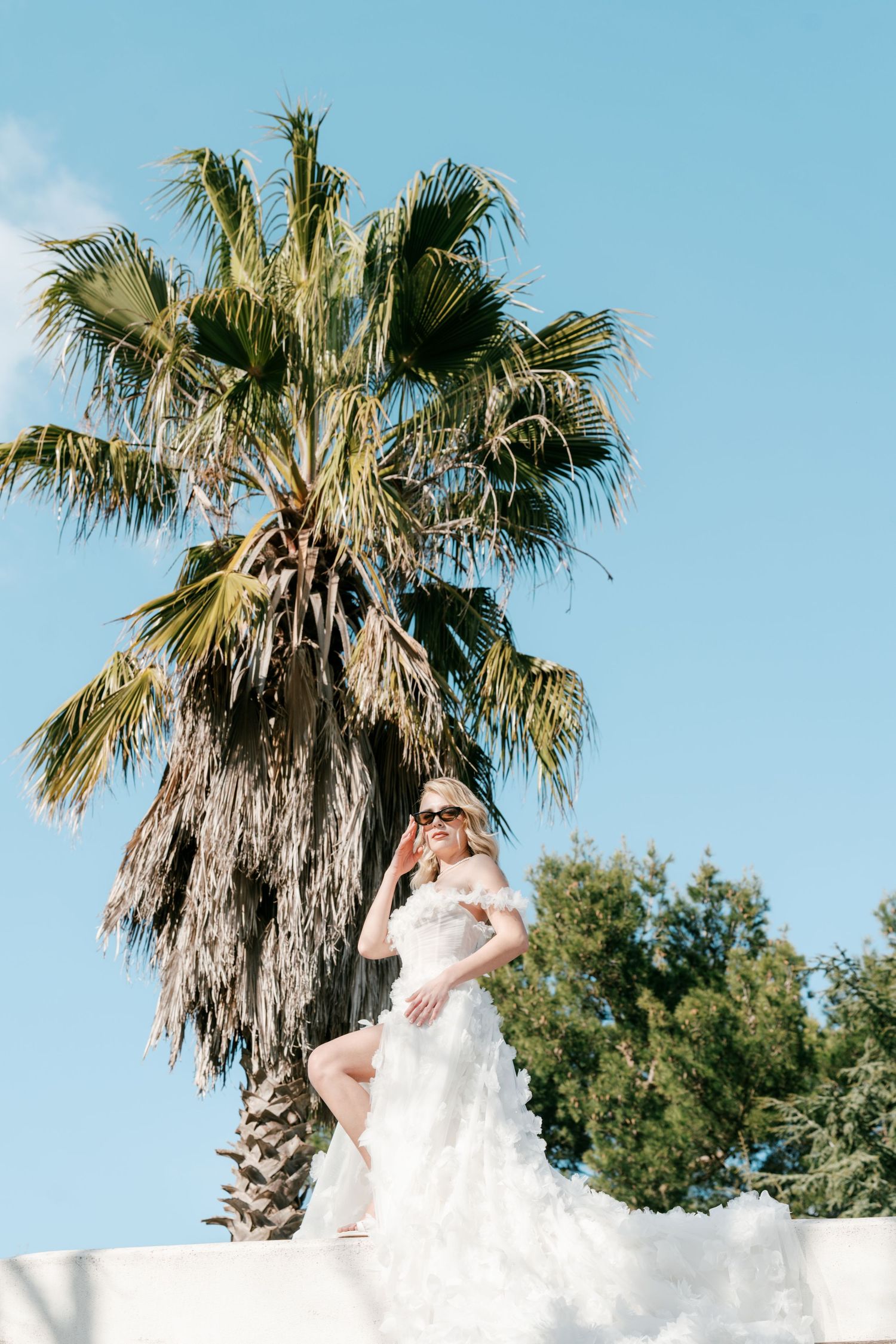 Blonde bride in white ruffled off-shoulder gown and sunglasses posing by a palm tree under blue sky. Mallorca wedding photographer, film