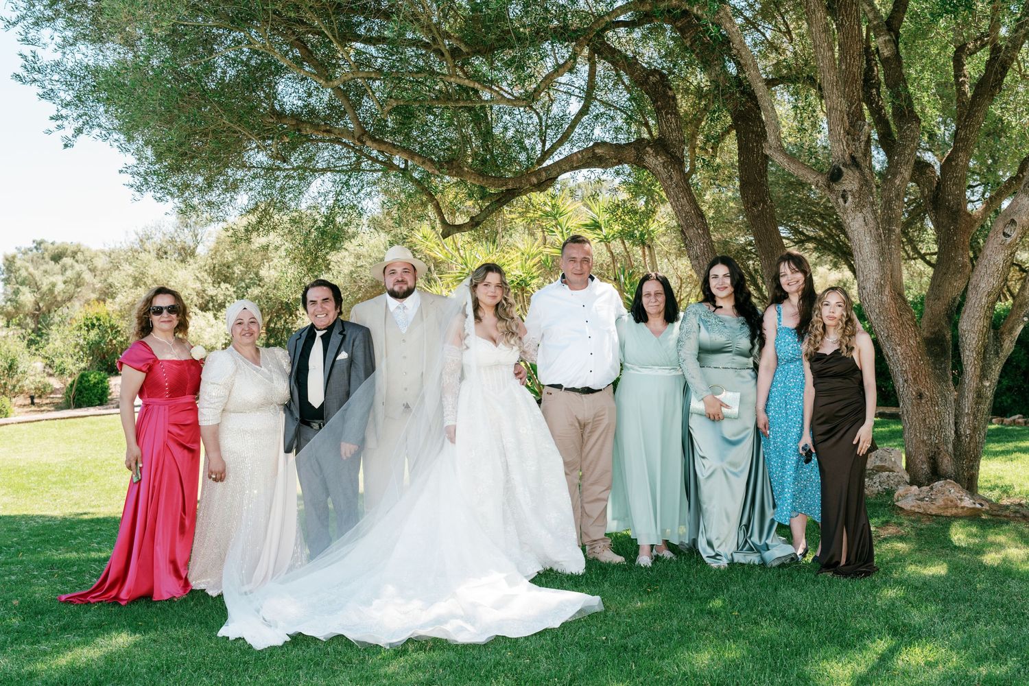 Wedding party posing outdoors under a large tree on a sunny day, bride in white gown surrounded by family in Mallorca.