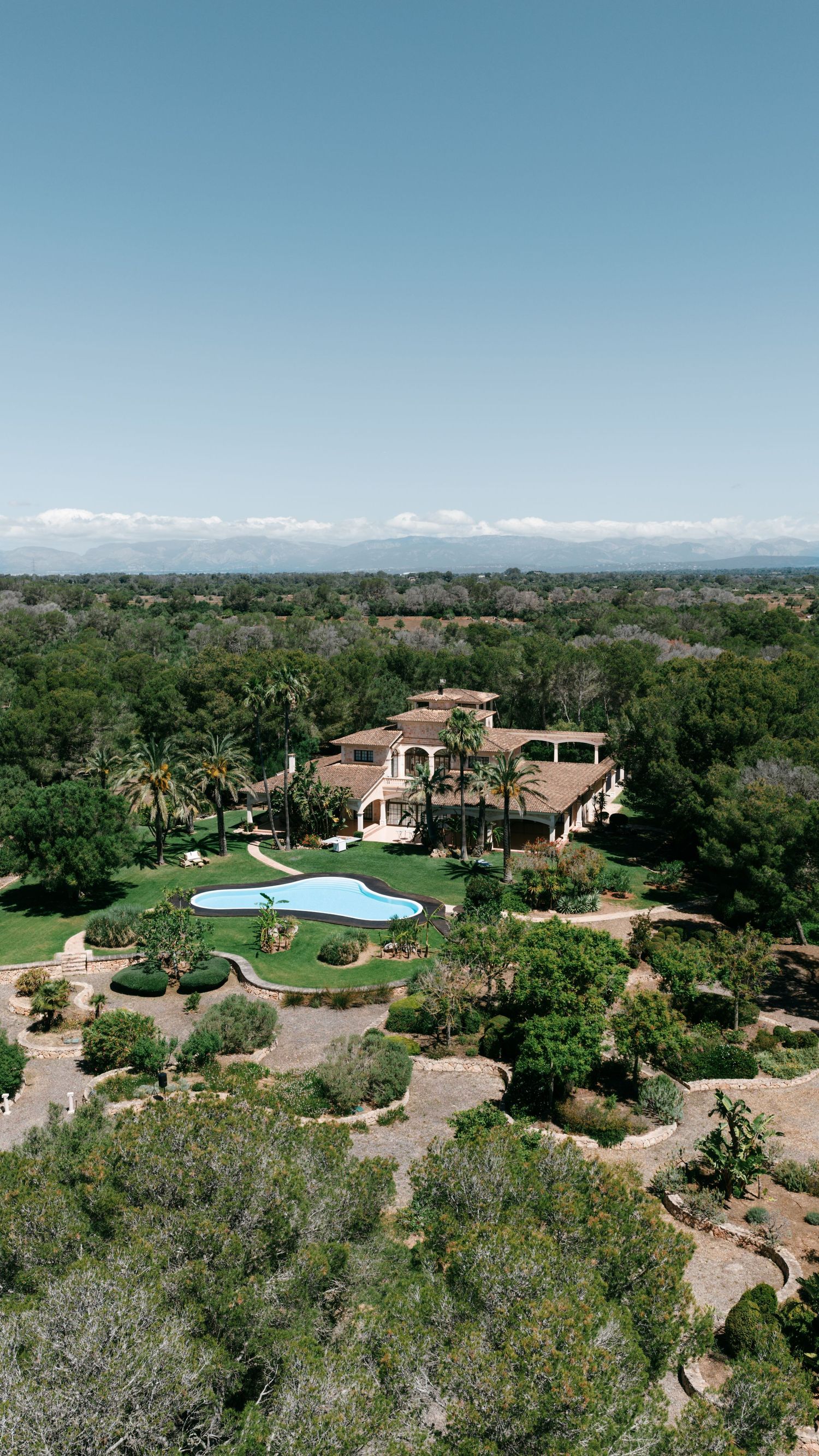 Aerial view of a luxury Mediterranean villa with a pool, manicured gardens, and palm trees surrounded by lush greenery.