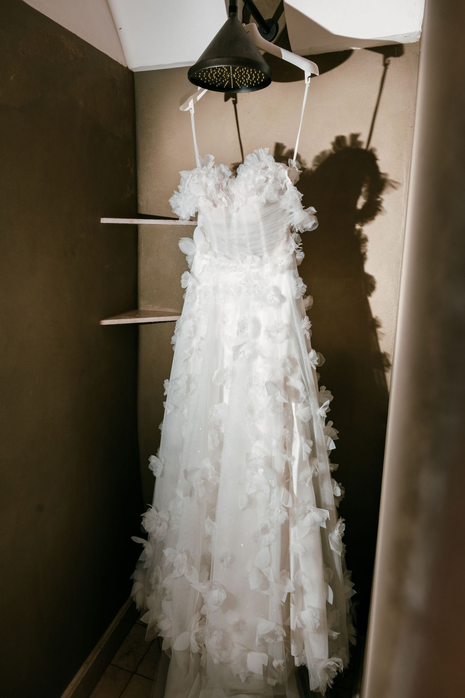 White floral applique wedding dress hanging on a shower head in a dimly lit room Borgo Stomennano.