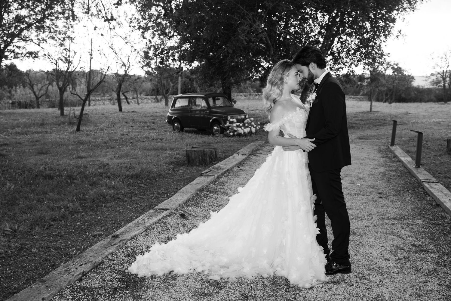 Black and white wedding photo of bride in ruffled gown and groom embracing outdoors near vintage car and trees.