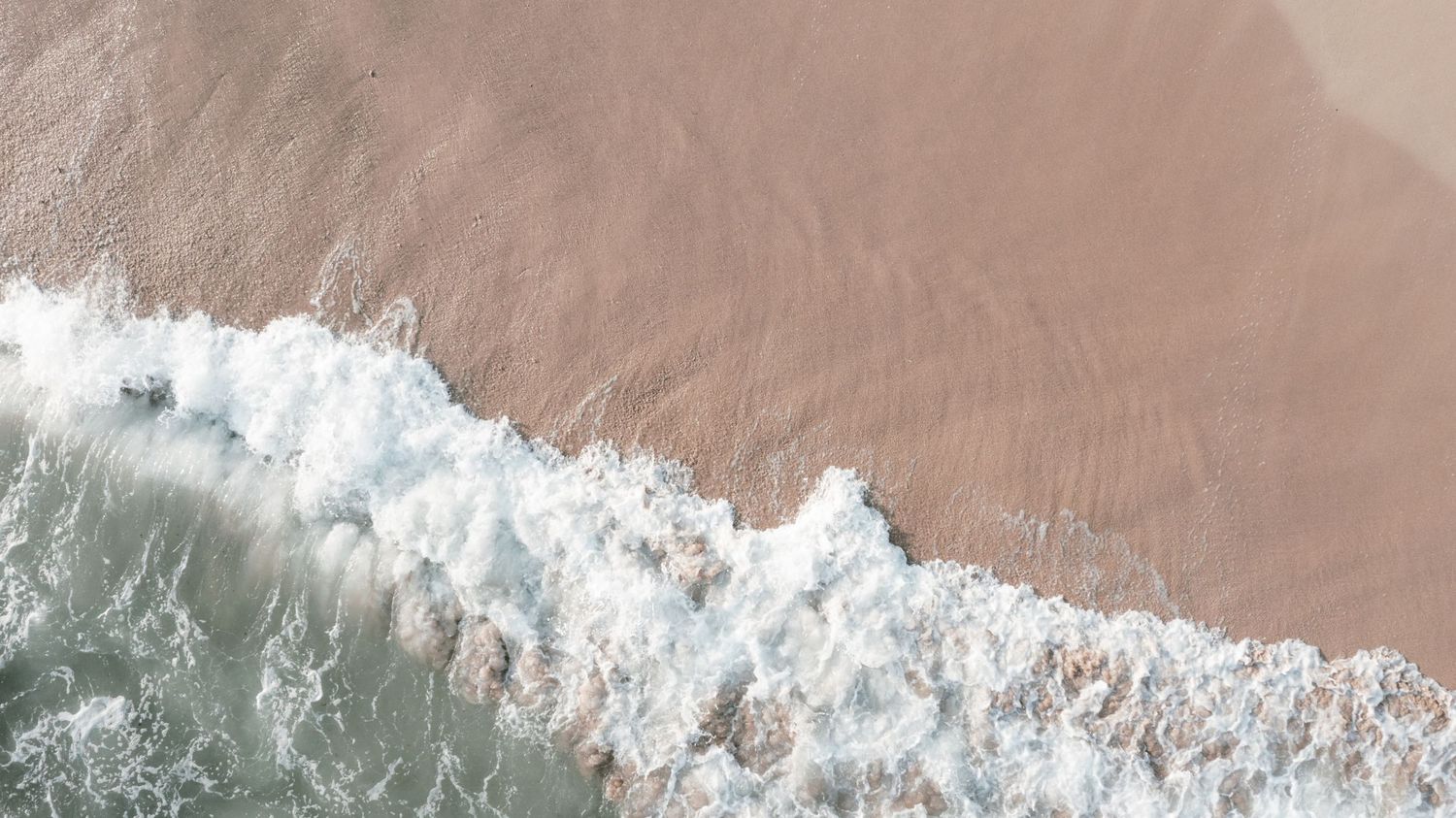 Aerial view of ocean waves crashing onto a sandy beach shoreline with white sea foam. Mallorca wedding photographer.