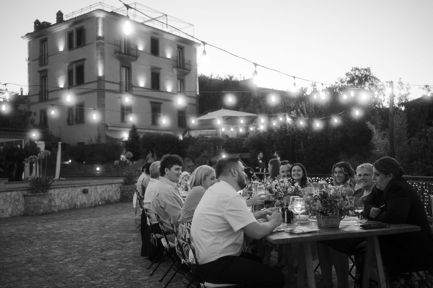 Wedding guests dining at long outdoor table under string lights with illuminated white villa in black and white. Tuscany wedding photographe