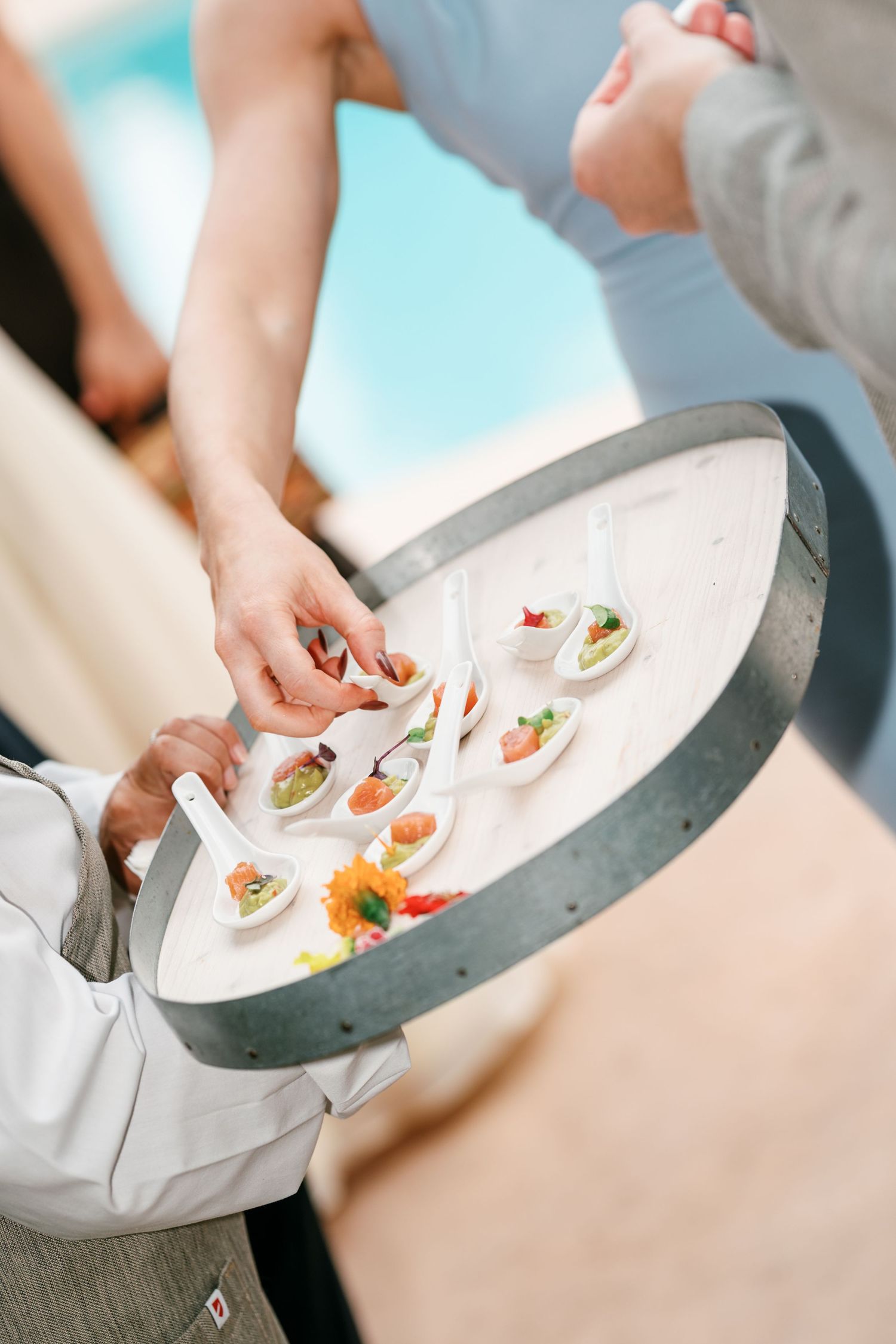 A guest picks up a ceramic spoon of gourmet hors d'oeuvres from a server's round wooden tray at an outdoor poolside event.