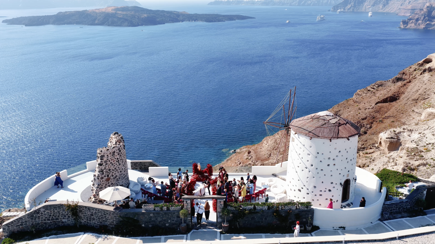 Aerial view of a cliffside wedding ceremony beside a whitewashed windmill overlooking the deep blue Aegean Sea in Santorini, Greece.