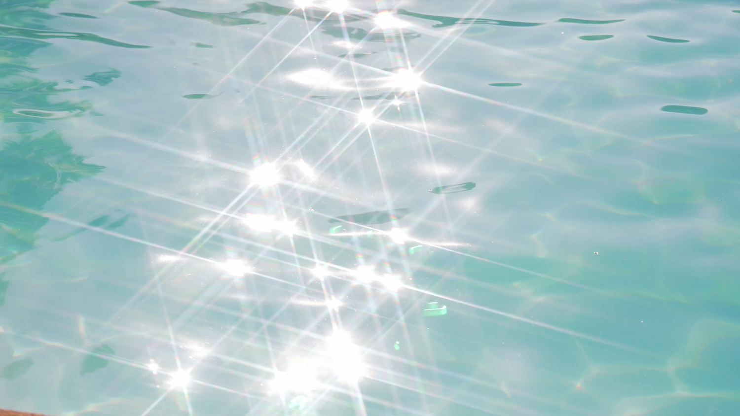 Bright sunlight sparkles on the rippling surface of clear turquoise swimming pool water, creating starburst glints.