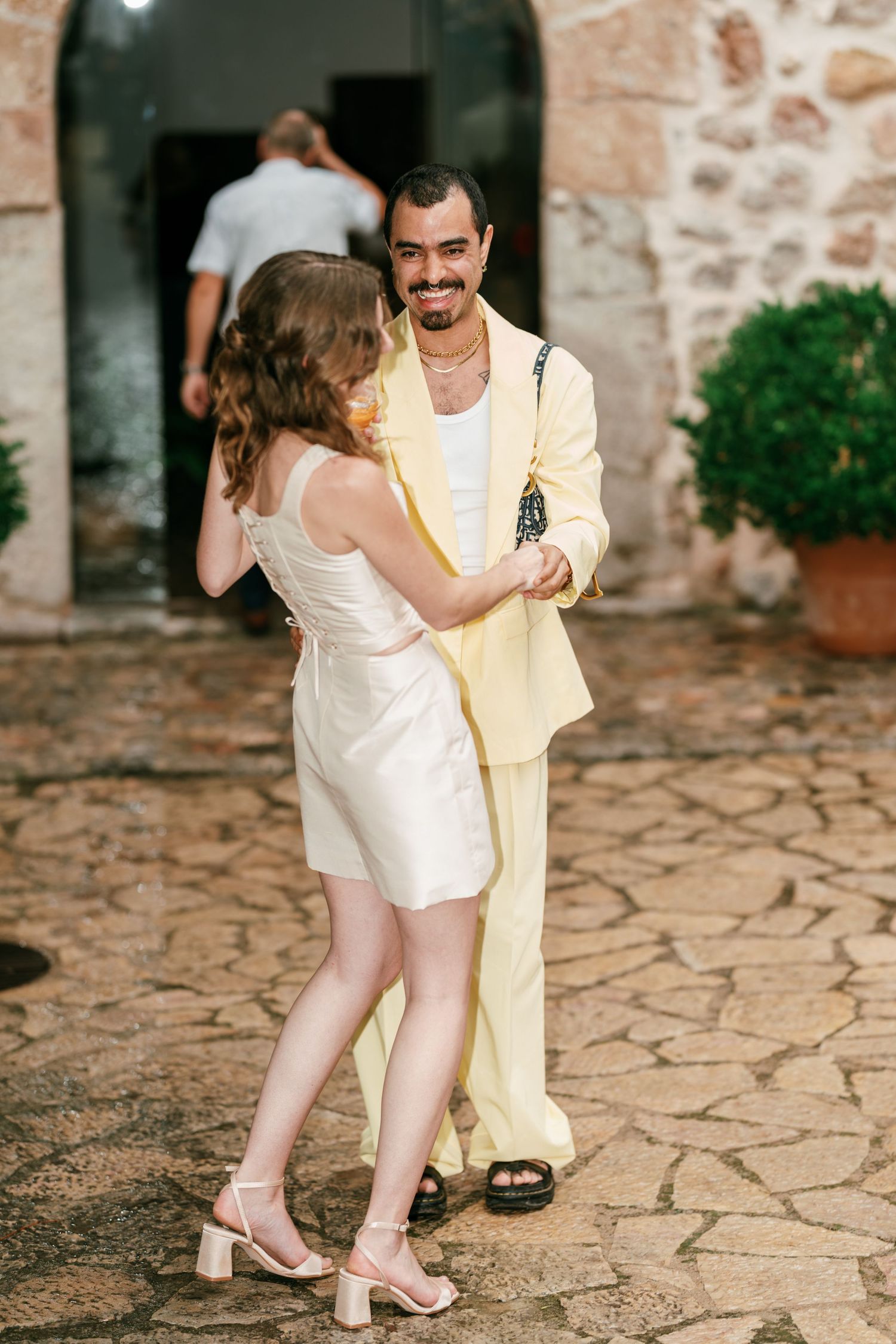 A couple in pastel yellow and cream outfits dances on a cobblestone courtyard beside a stone archway at an outdoor wedding celebration.