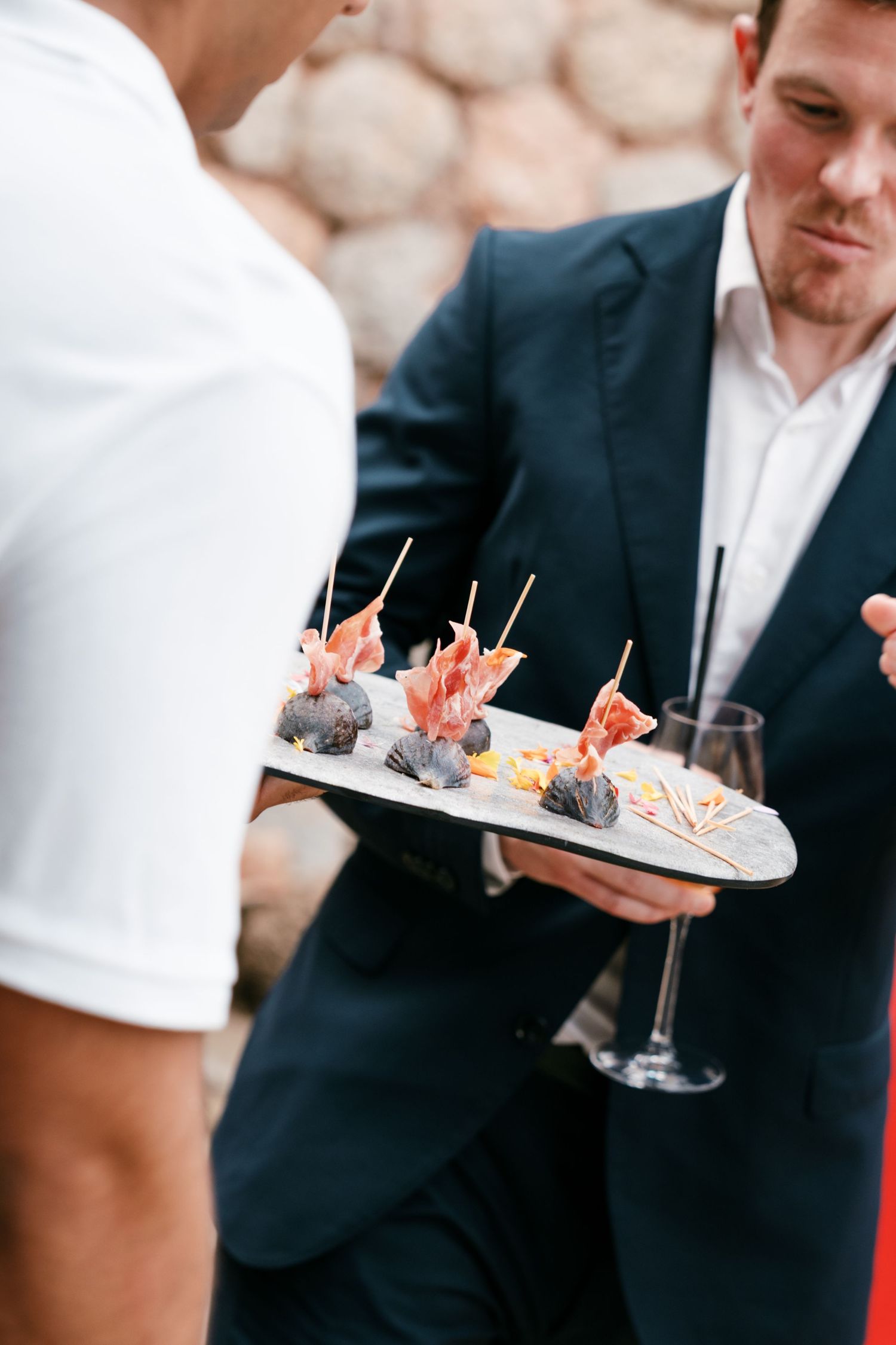 Man in a navy suit holds a slate serving tray of fig and prosciutto skewers and a wine glass at an outdoor catered reception.