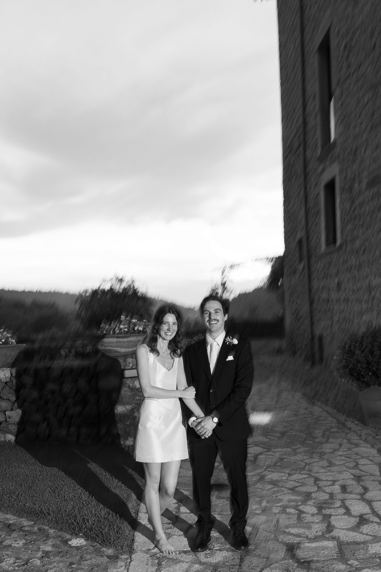 Black and white photo of a smiling couple holding hands on a cobblestone path, the bride in a short white dress and groom in a dark suit.